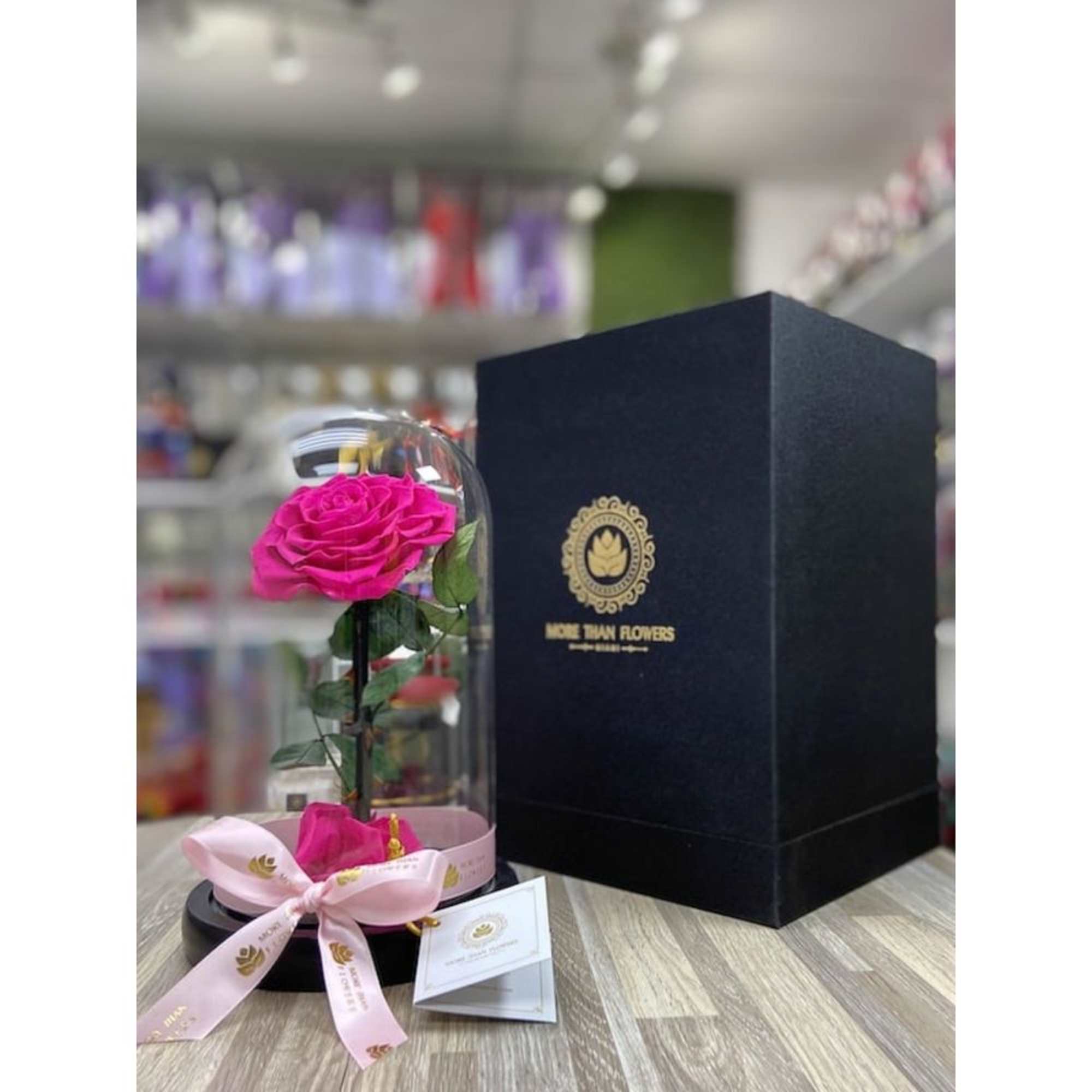 Hot pink rose in a glass dome on a black base with ribbon, next to a tall black gift box