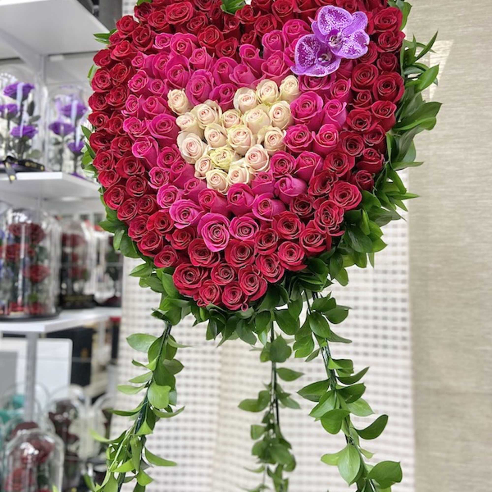 Tall heart-shaped arrangement of pink and red roses with cream center and trailing greenery on a stand