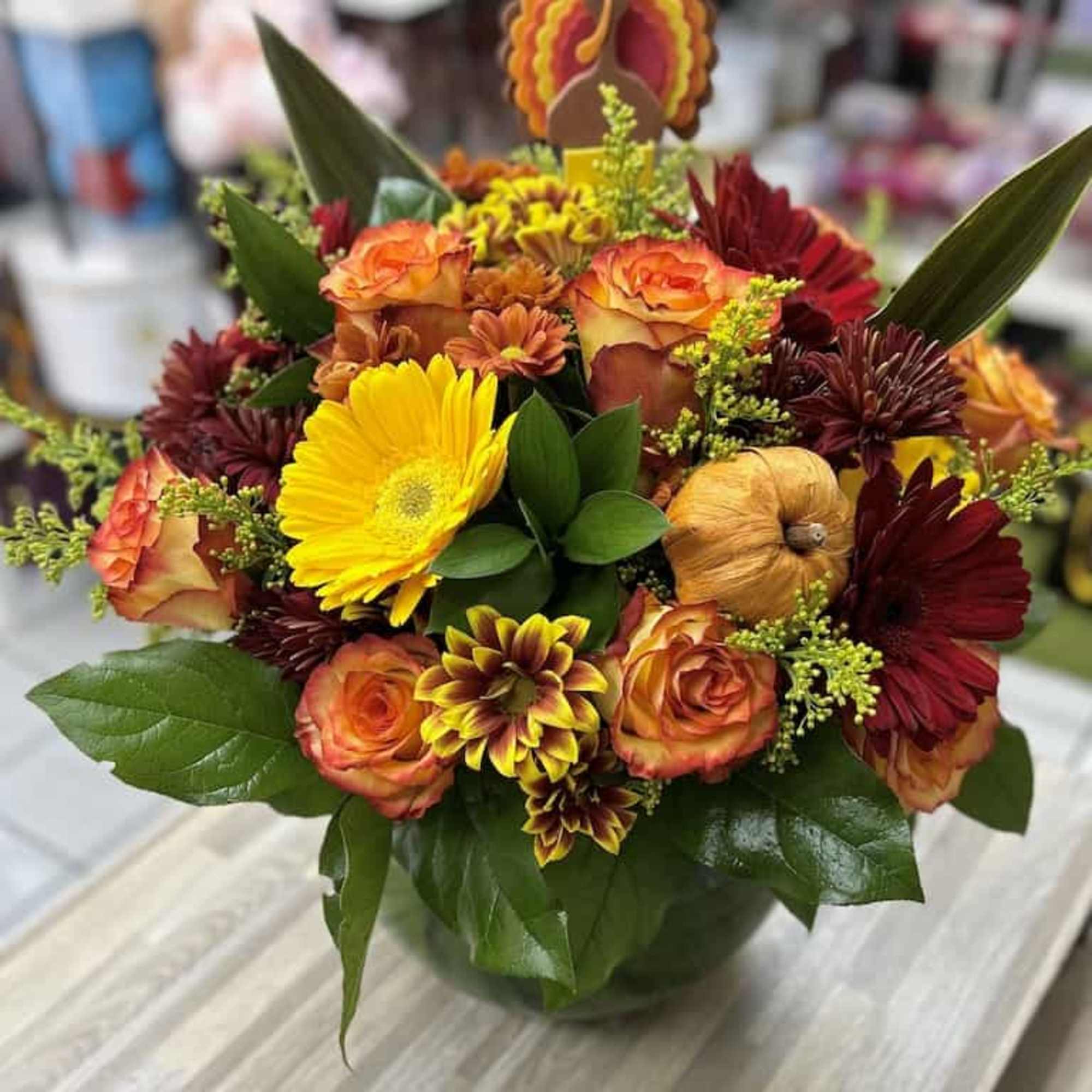 Round arrangement of orange roses, yellow gerbera, and red mums in a glass bowl with a turkey pick.