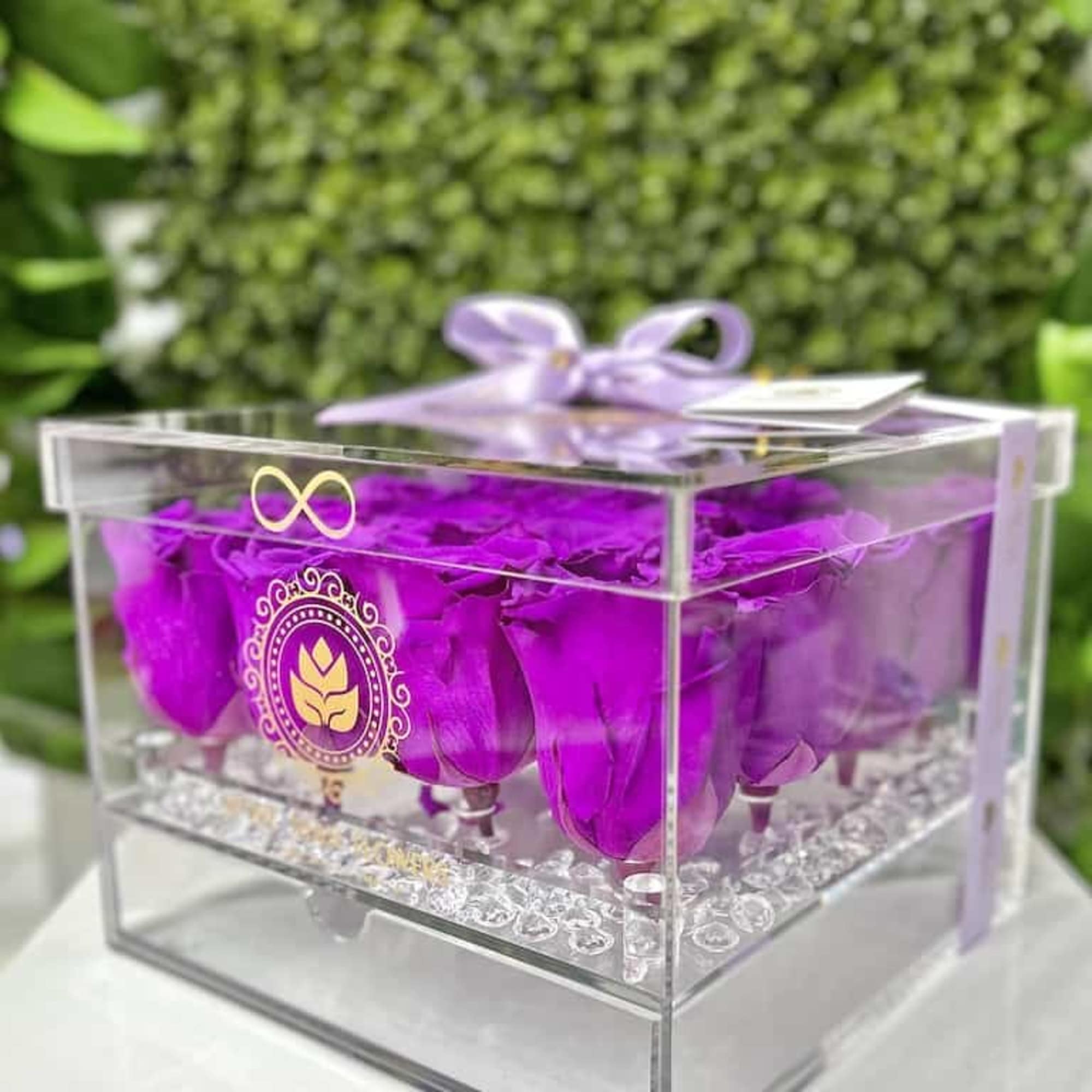 Clear acrylic box filled with bright purple roses and topped with a lavender ribbon bow