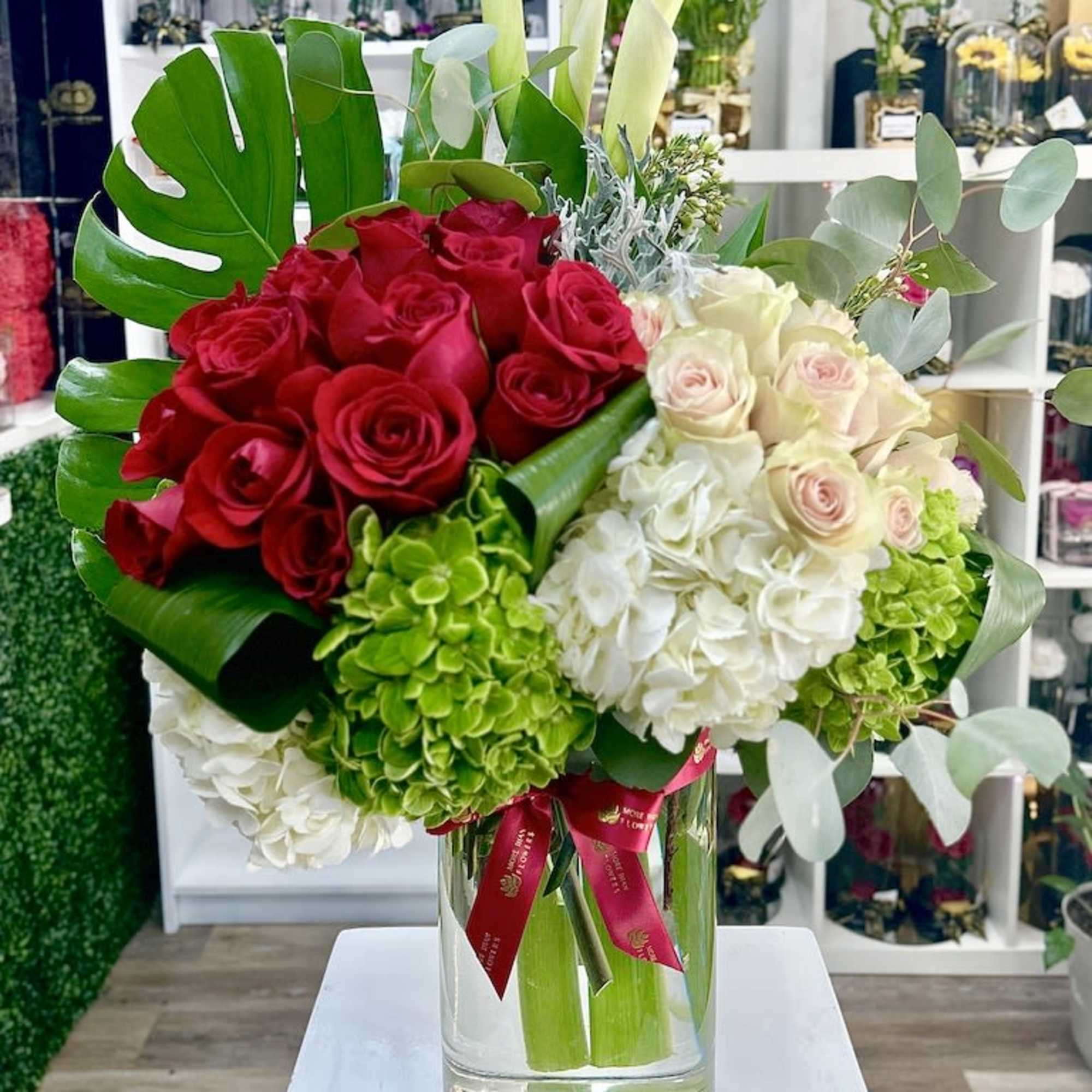 Tall glass vase with red roses, white and green hydrangeas, calla lilies, and a large monstera leaf.