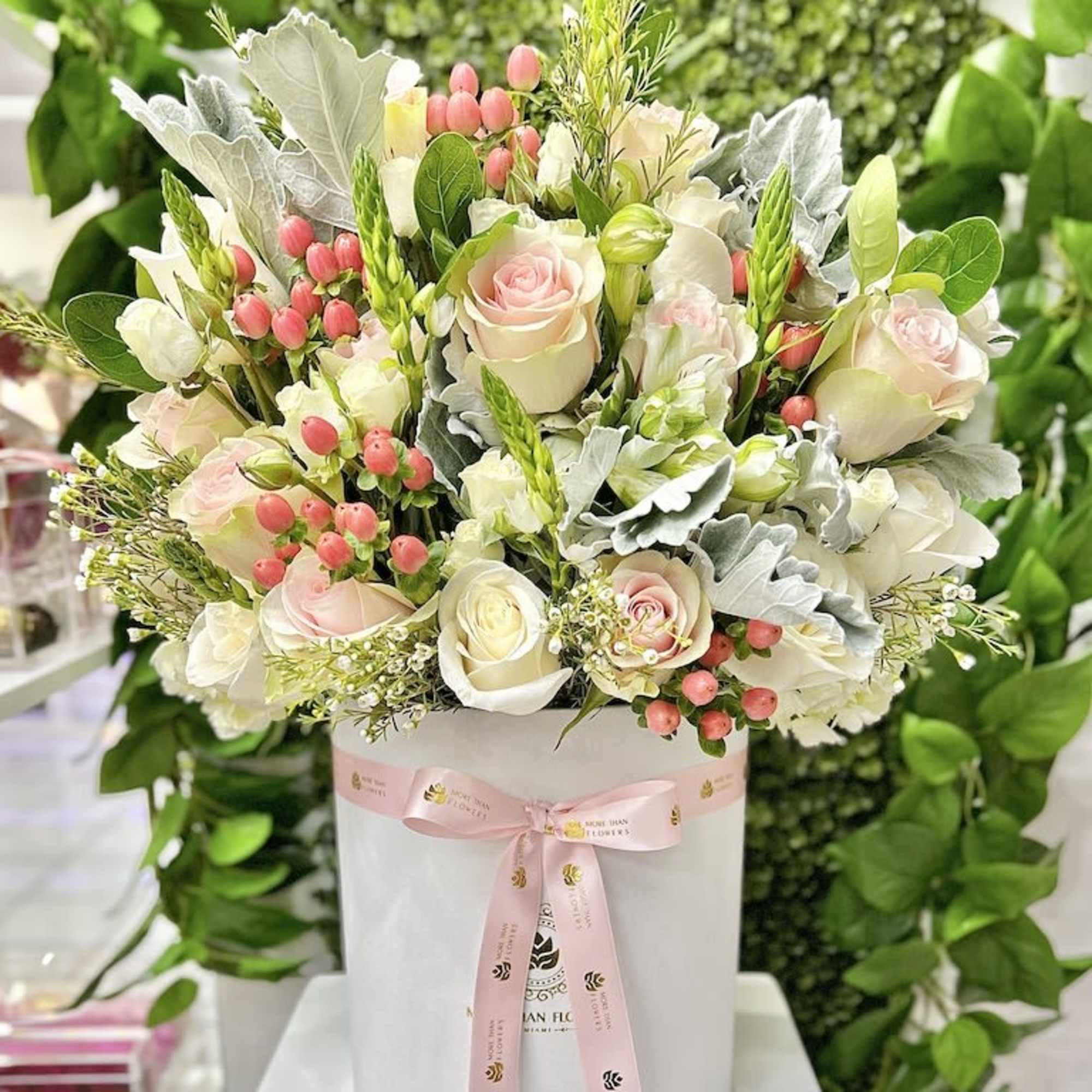Round hatbox arrangement of pale pink and white roses with pink berries and foliage, tied with a pink ribbon.