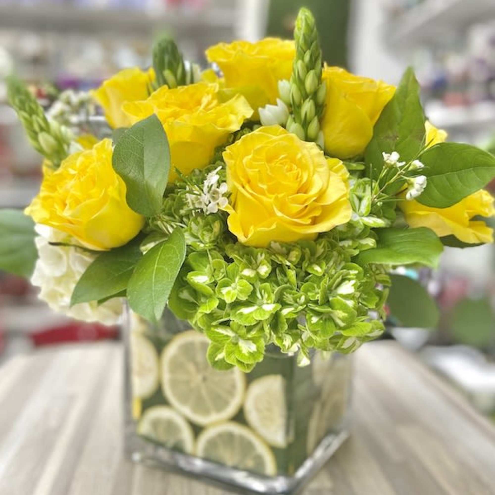 Square glass vase filled with lemon slices holding yellow roses and green hydrangeas with white accents.
