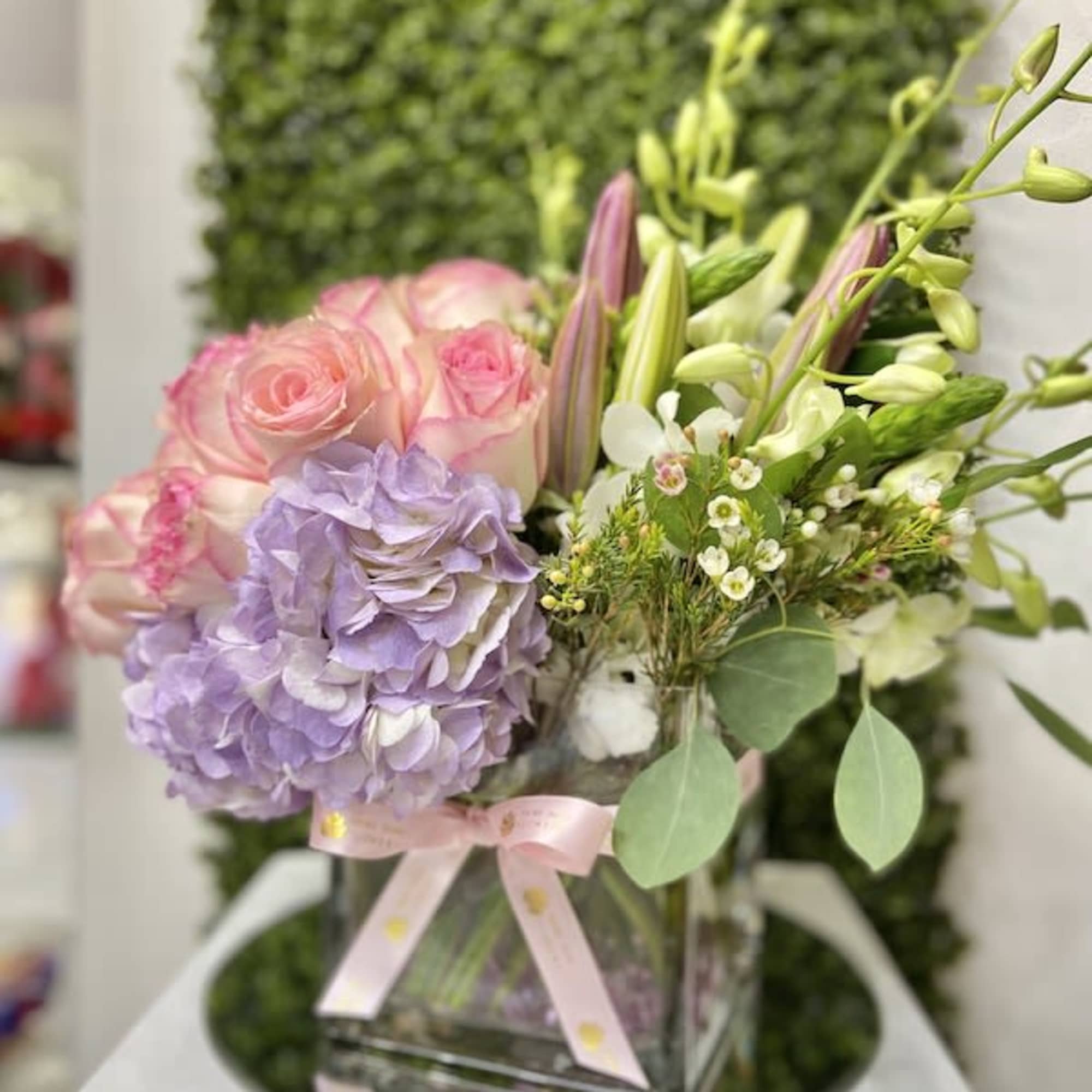 Pastel arrangement of pink roses, lavender hydrangea, and lilies in a glass cube vase with a pink ribbon.