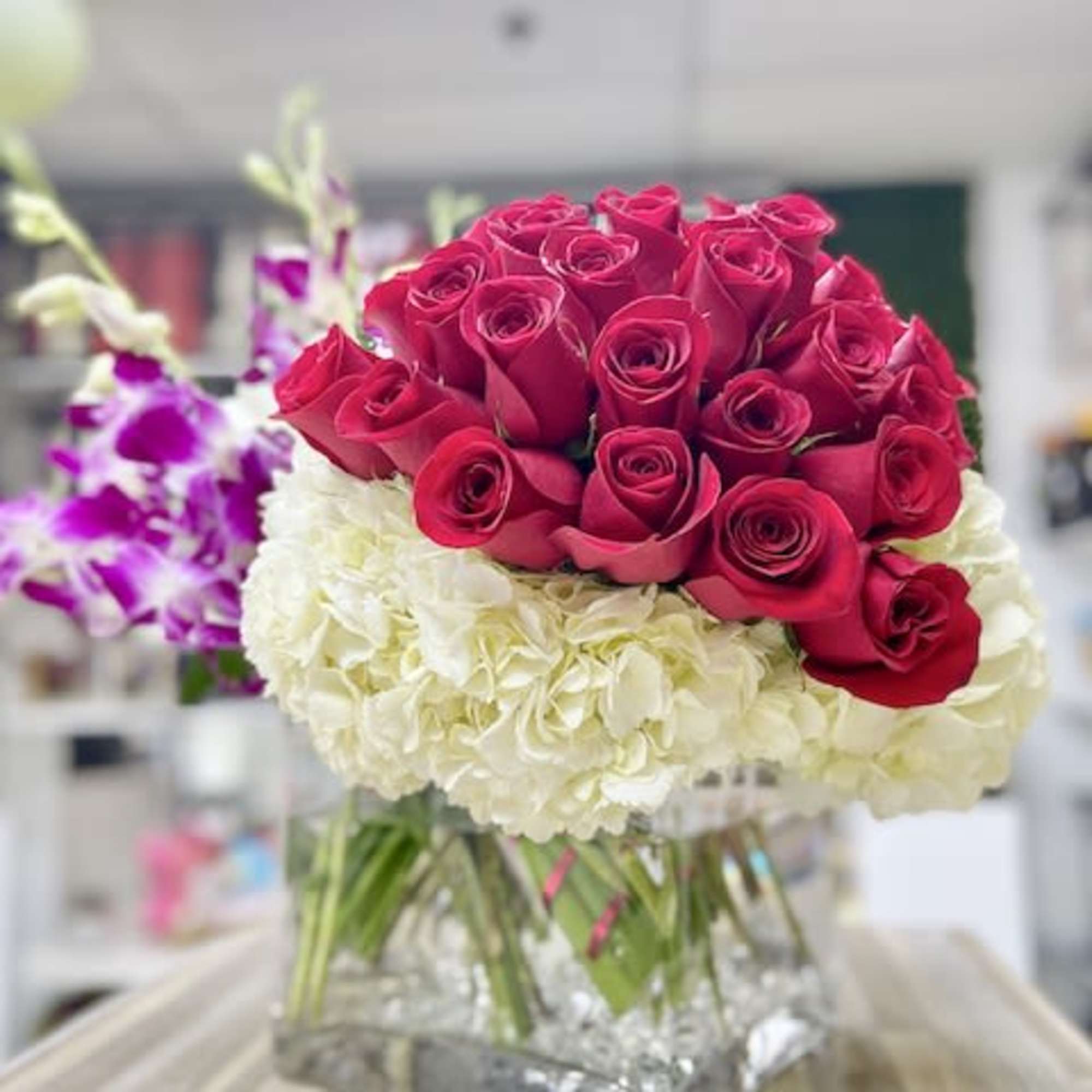 Red roses arranged over white hydrangeas with purple orchids in a clear square glass vase