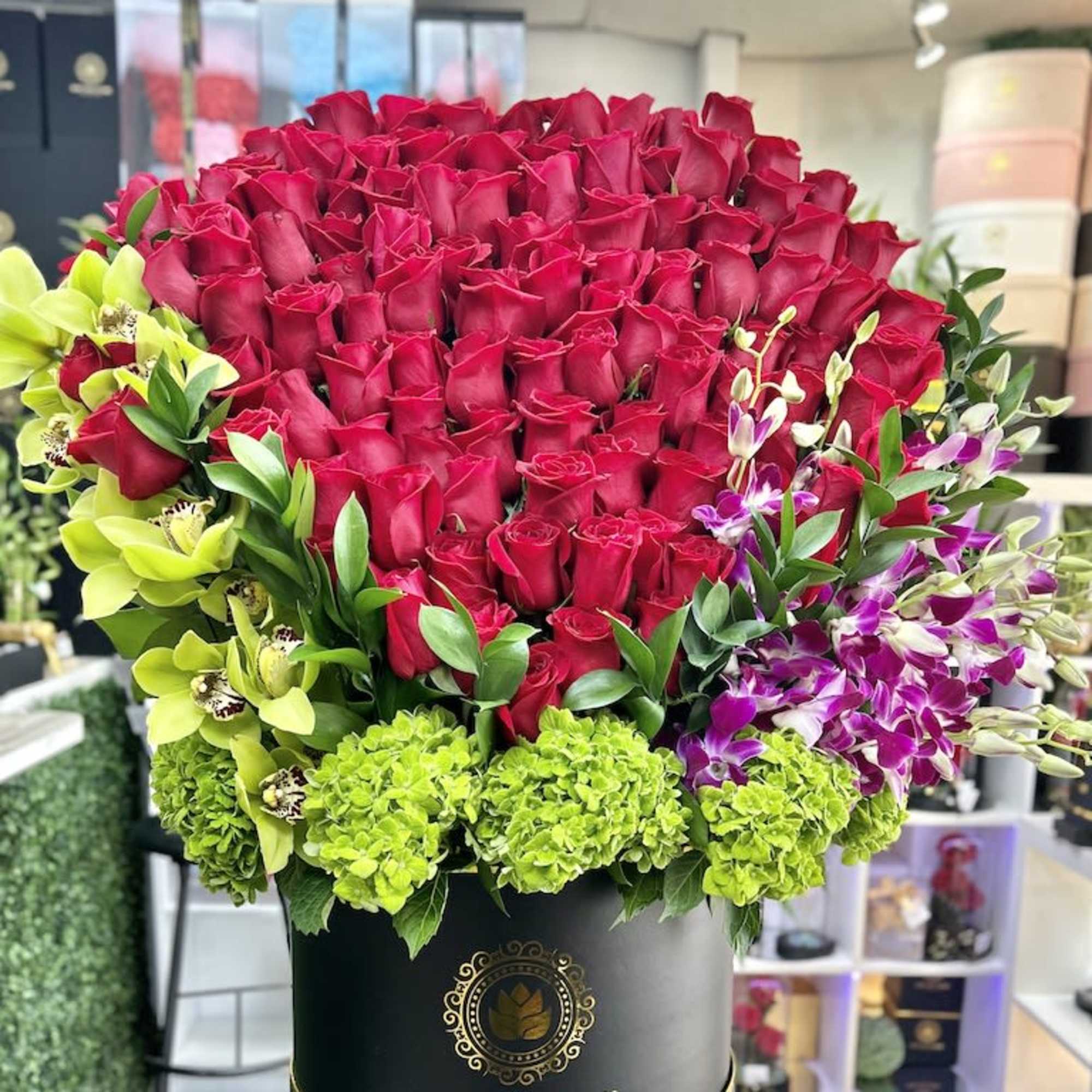 Large round box overflowing with red roses, green hydrangeas, and orchids in bright green and purple tones.