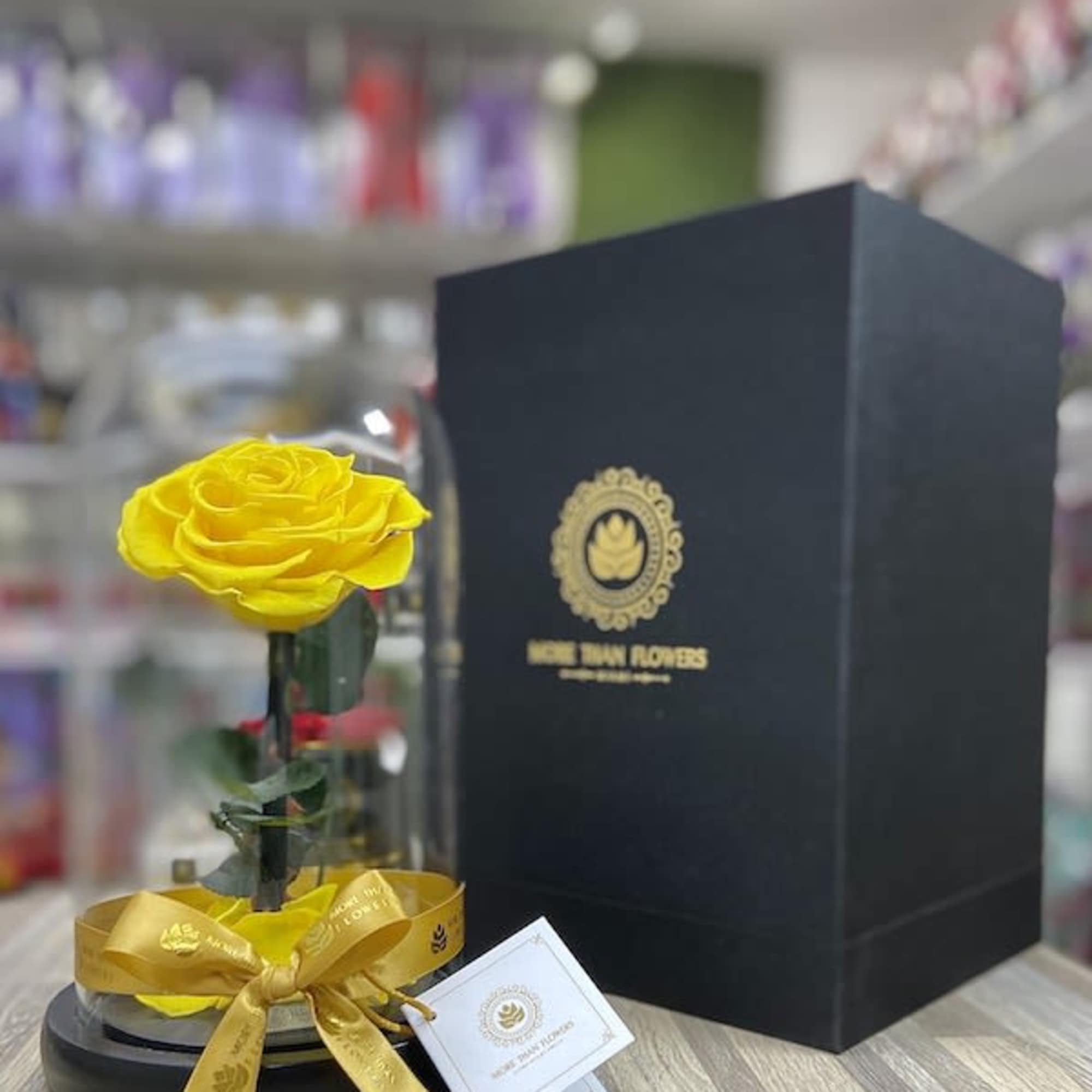 Single yellow rose under a glass dome with gold ribbon on black base beside a tall black gift box
