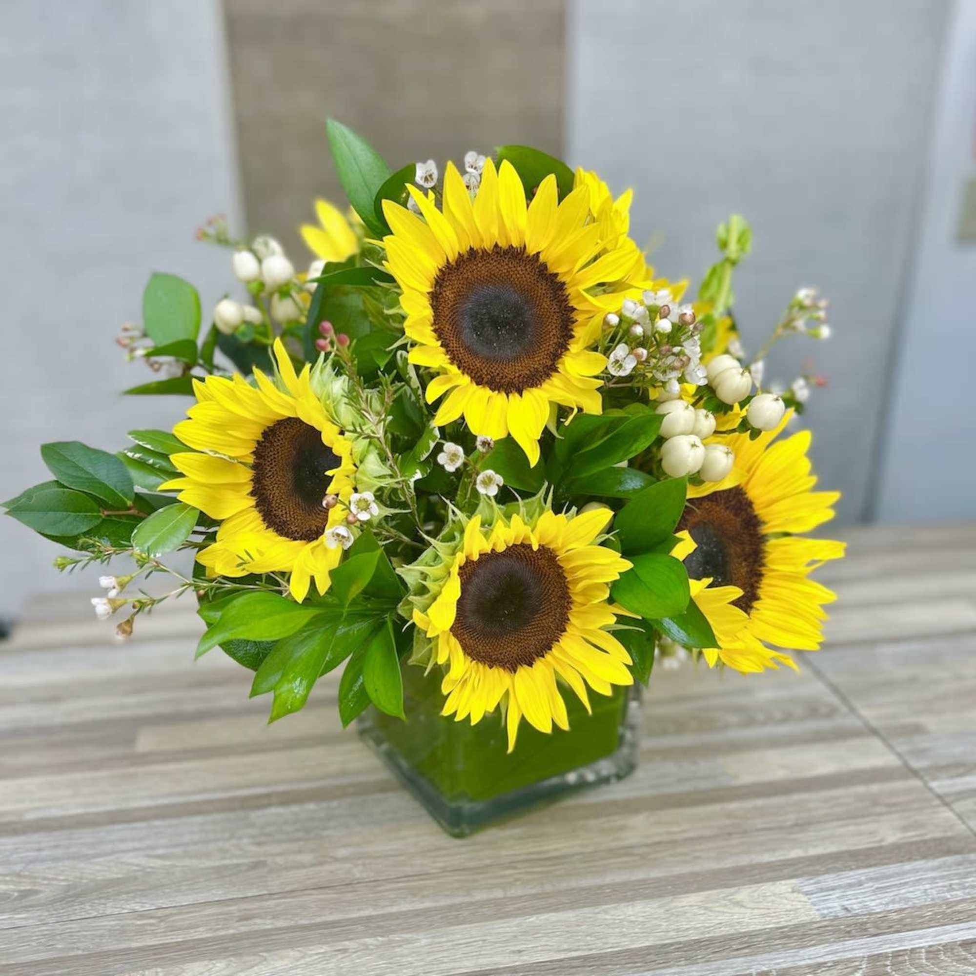 Arrangement of bright yellow sunflowers with small white blooms in a square glass vase.