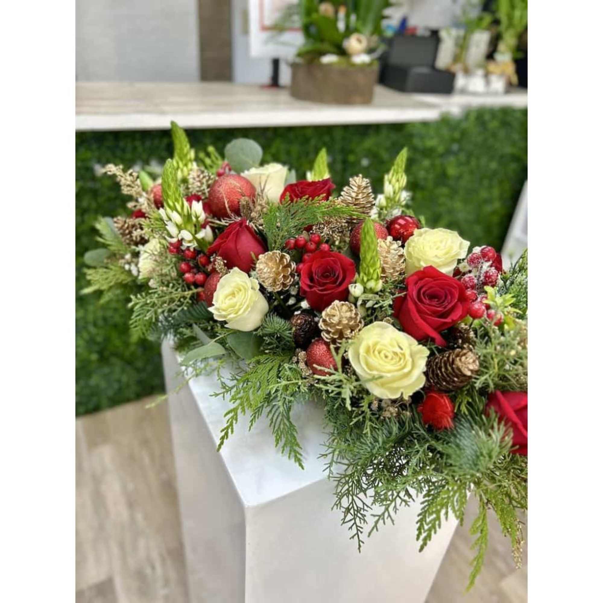 Long centerpiece of red and cream roses with evergreens, berries, and gold pinecones in a festive holiday design