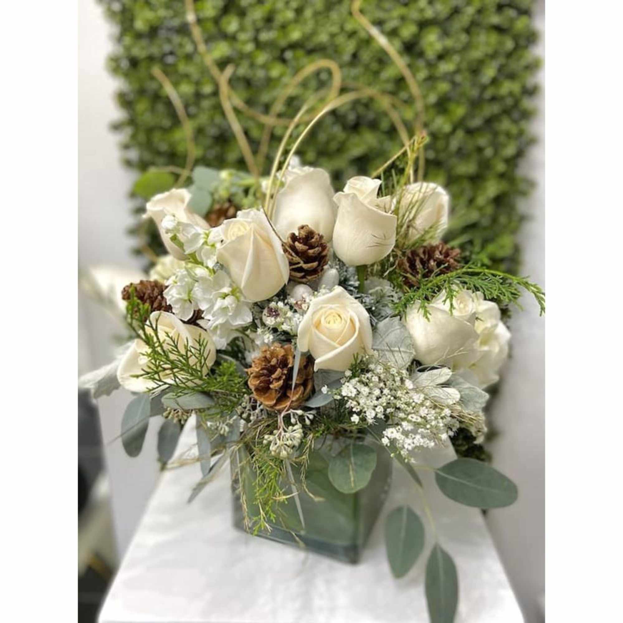 Winter arrangement of white roses with pinecones and evergreens in a square glass vase