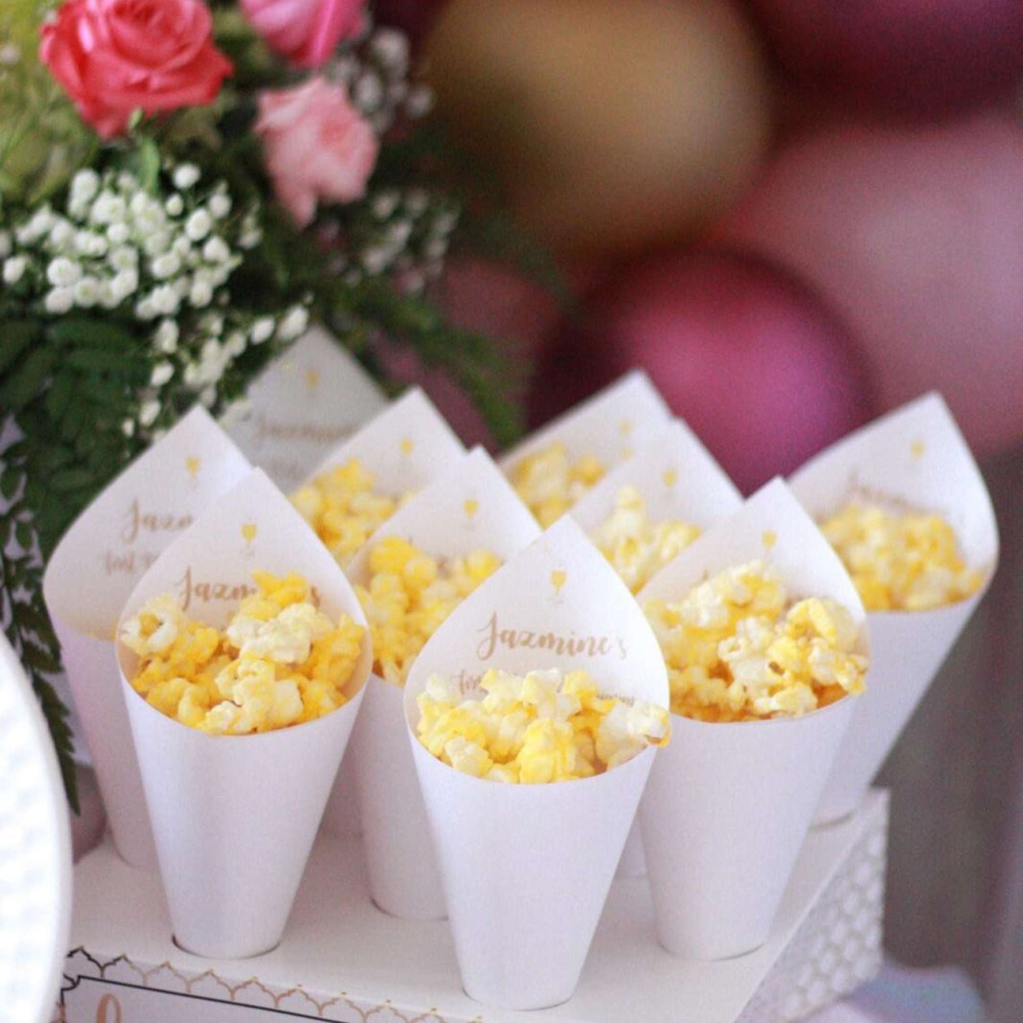 Popcorn favors in white cones on a decorated box with pink balloons and roses behind
