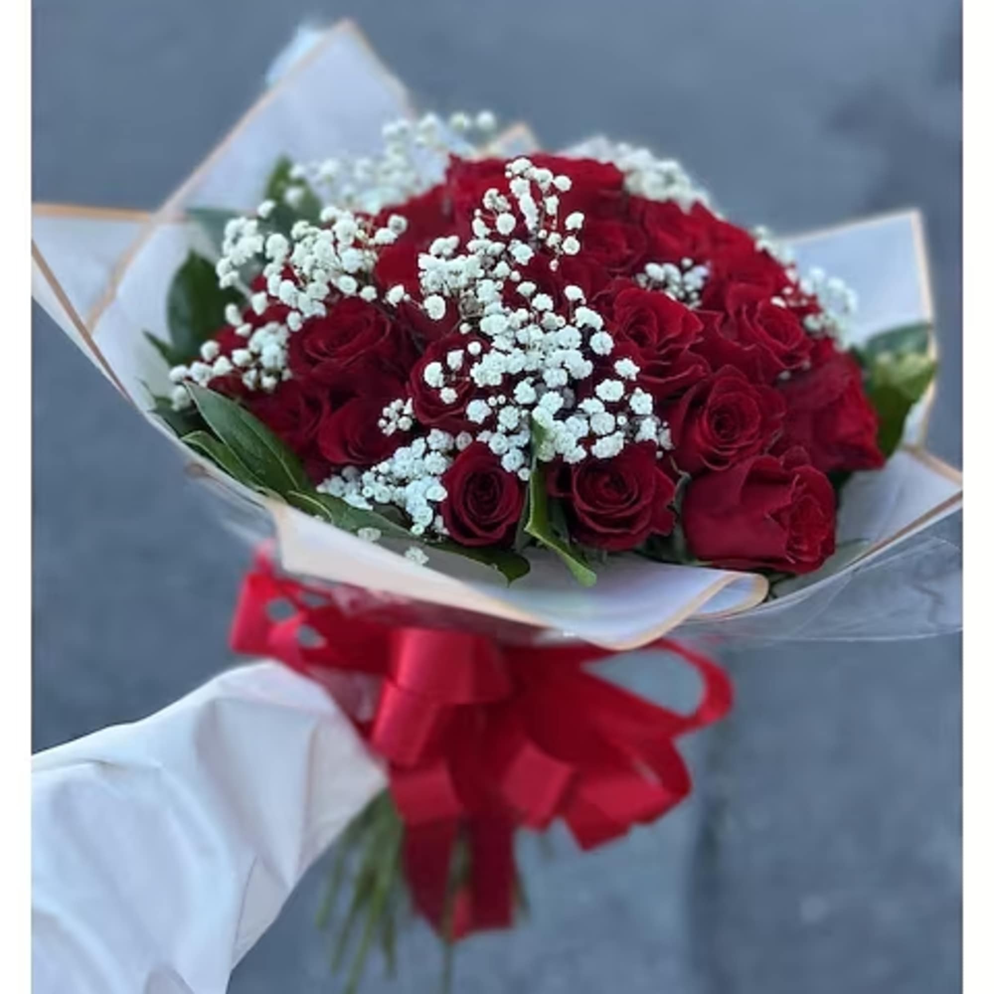 Handheld bouquet of red roses with white filler flowers wrapped in paper and tied with a large red bow.