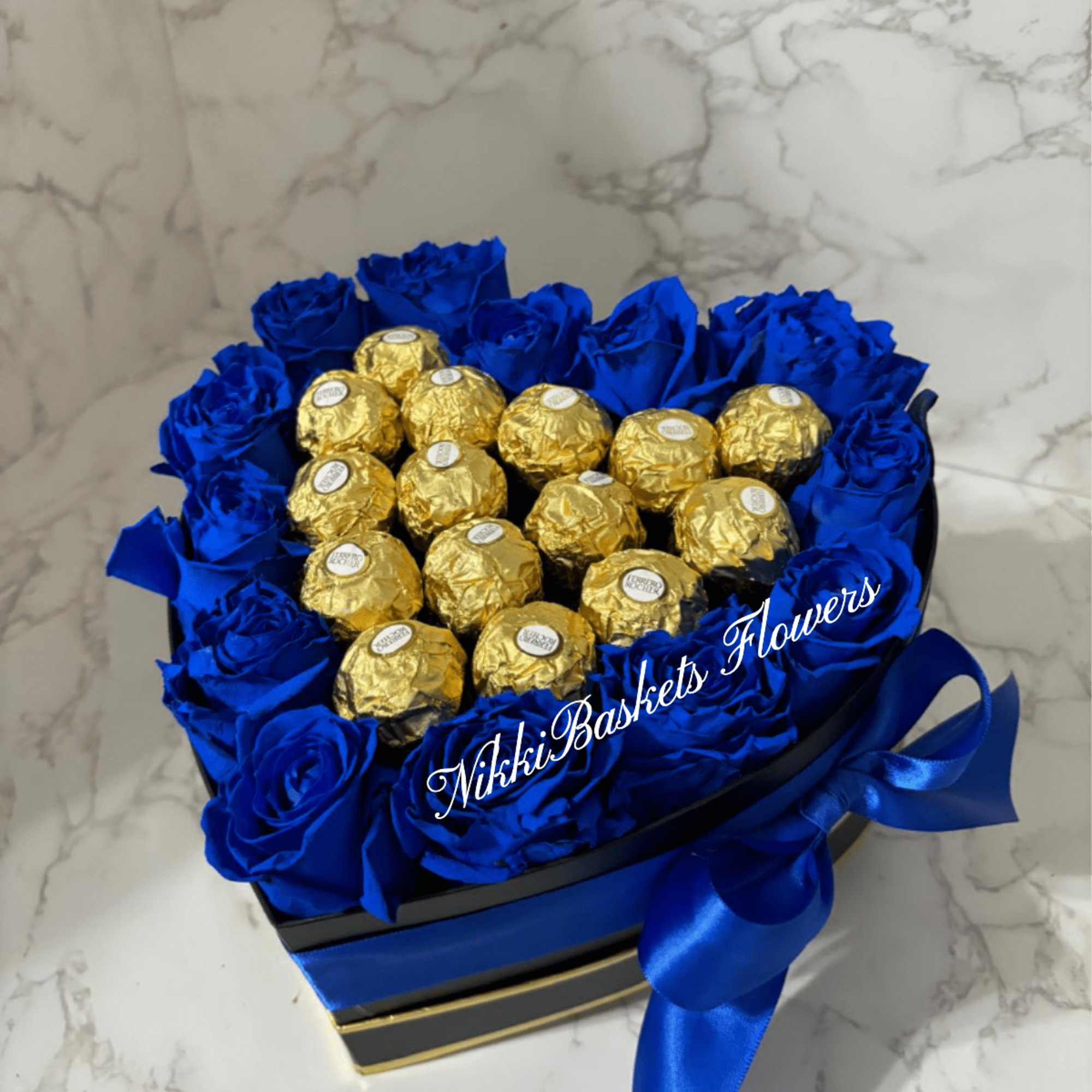 Blue roses arranged around gold-wrapped chocolates in a heart-shaped box