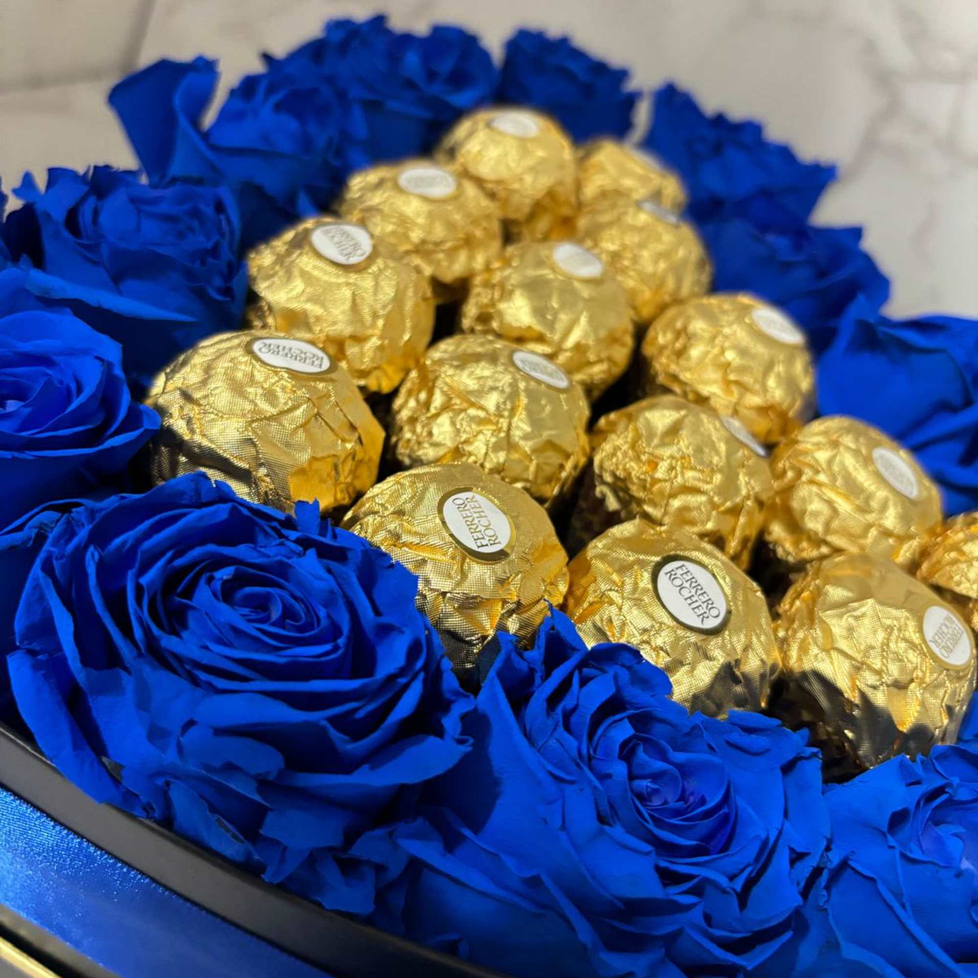 Blue roses arranged around gold-wrapped chocolates in a round box