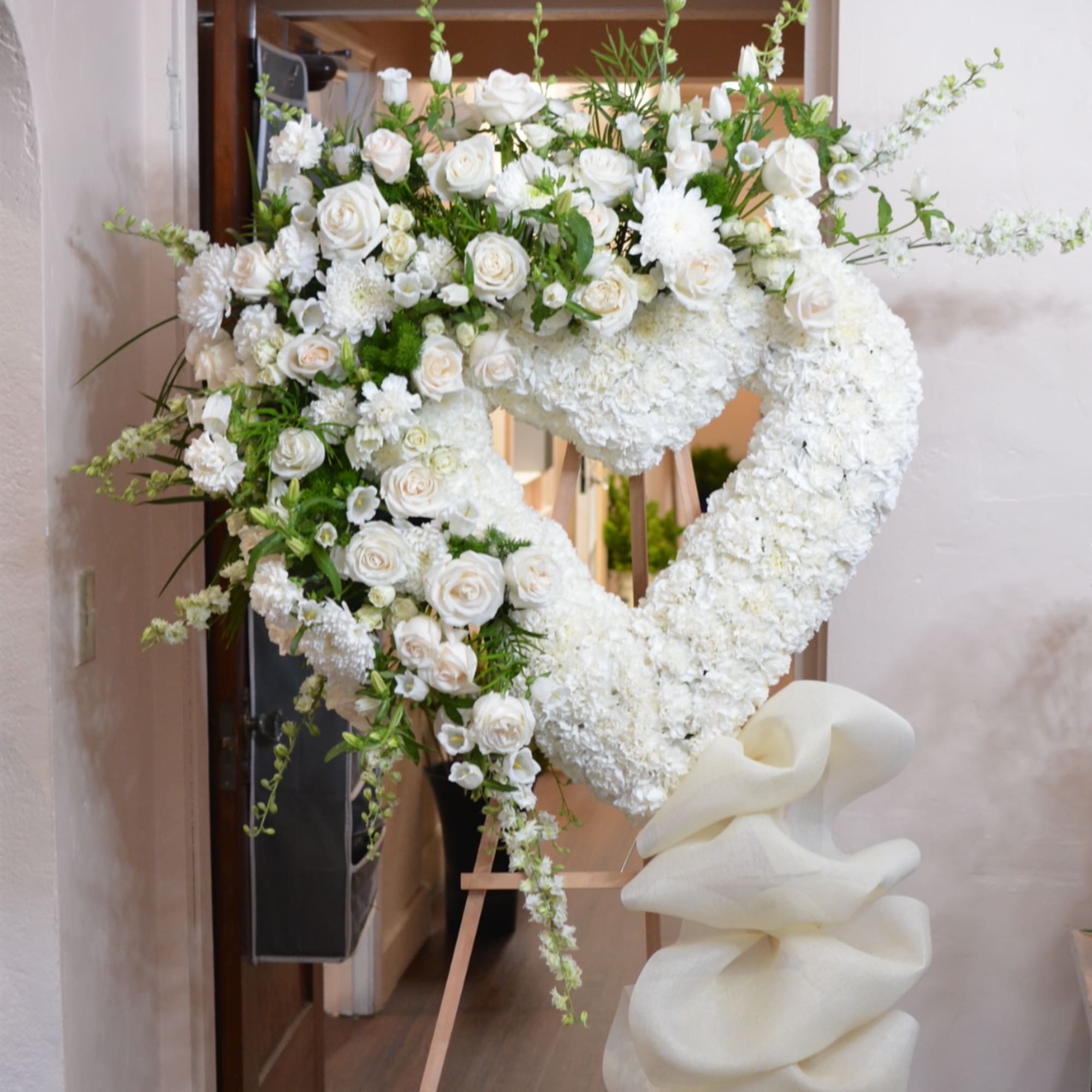 Heart-shaped white floral standing arrangement on an easel with a large white bow