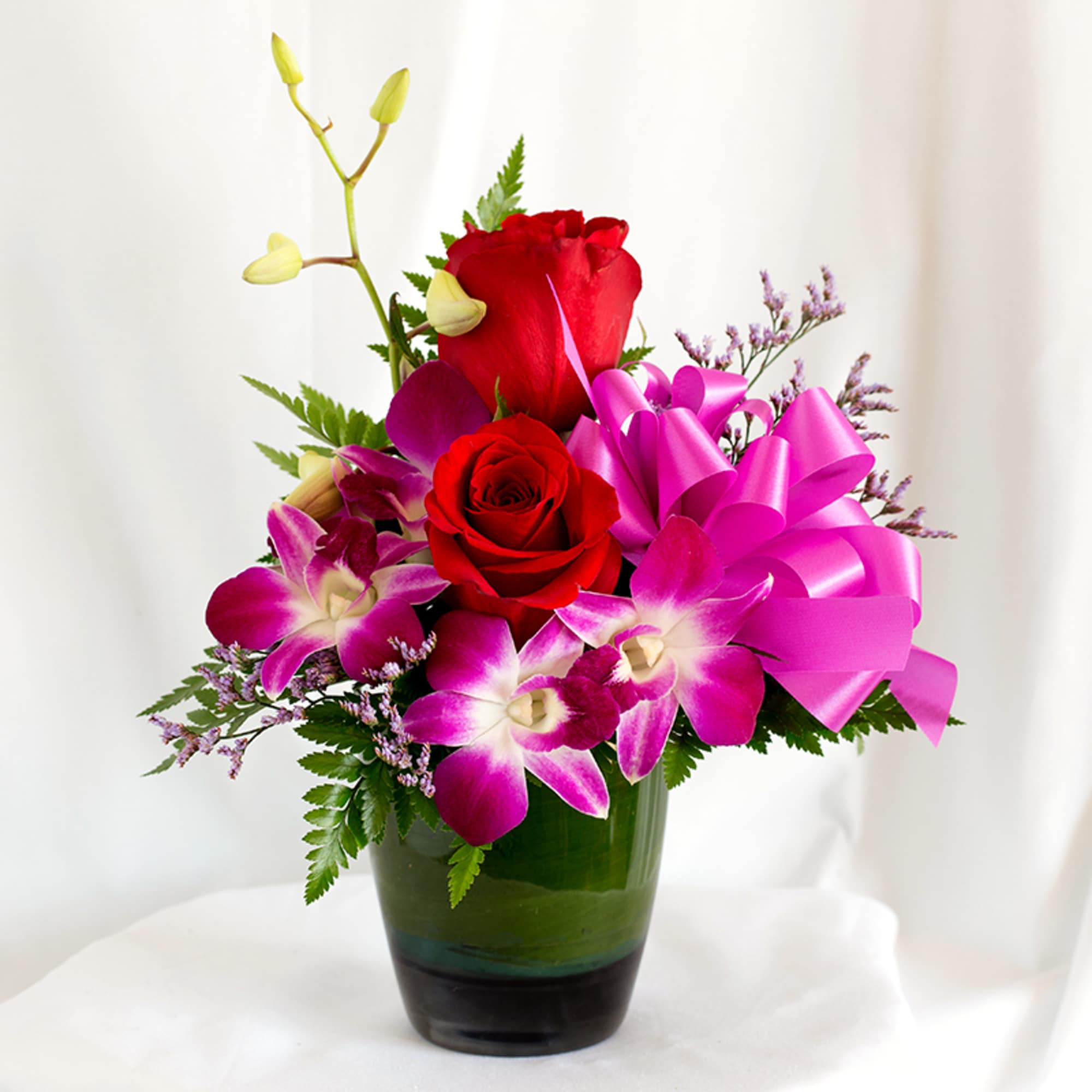 Red roses and purple orchids in a green glass vase with a pink ribbon