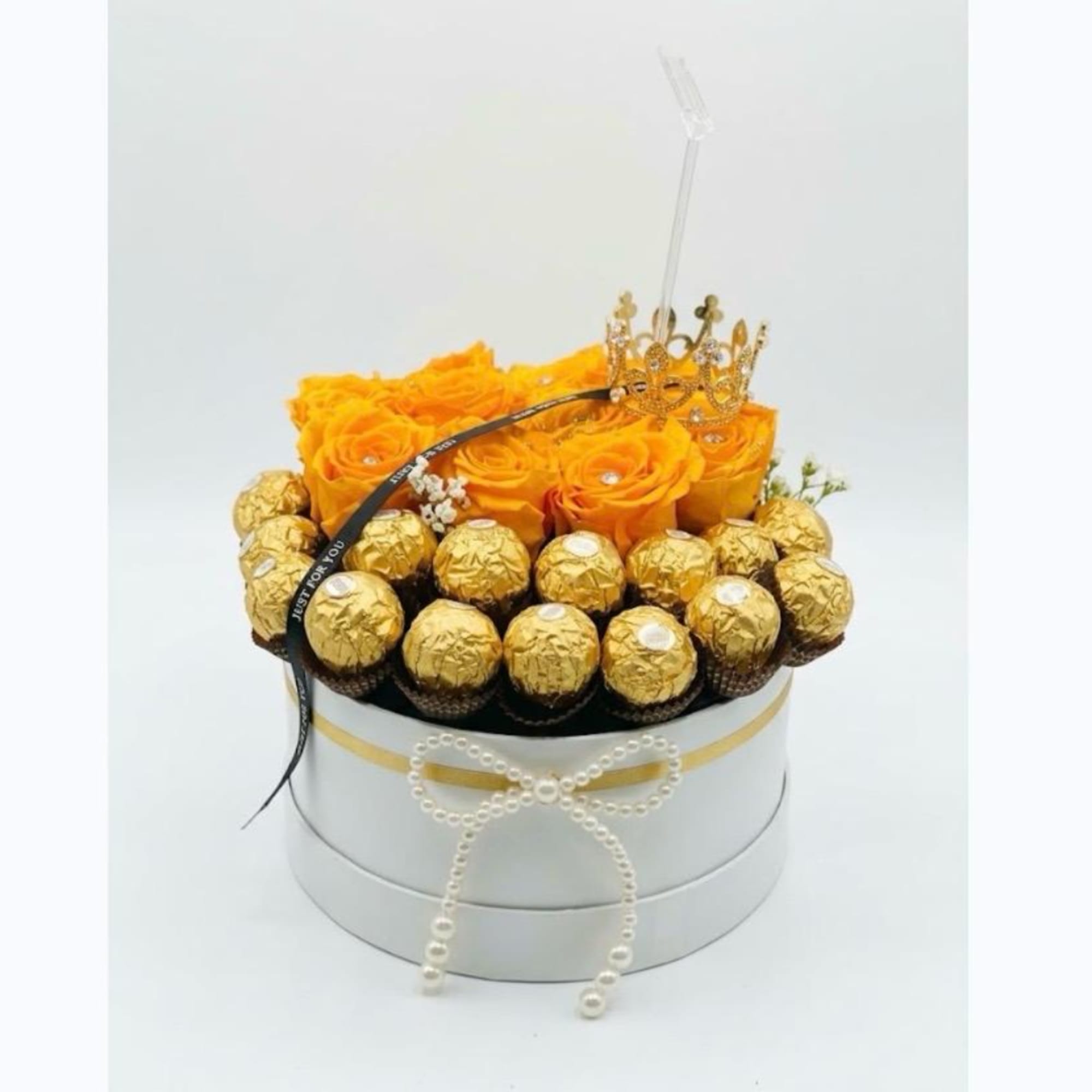 Orange roses in a round gift box with gold-wrapped chocolates and a crown topper