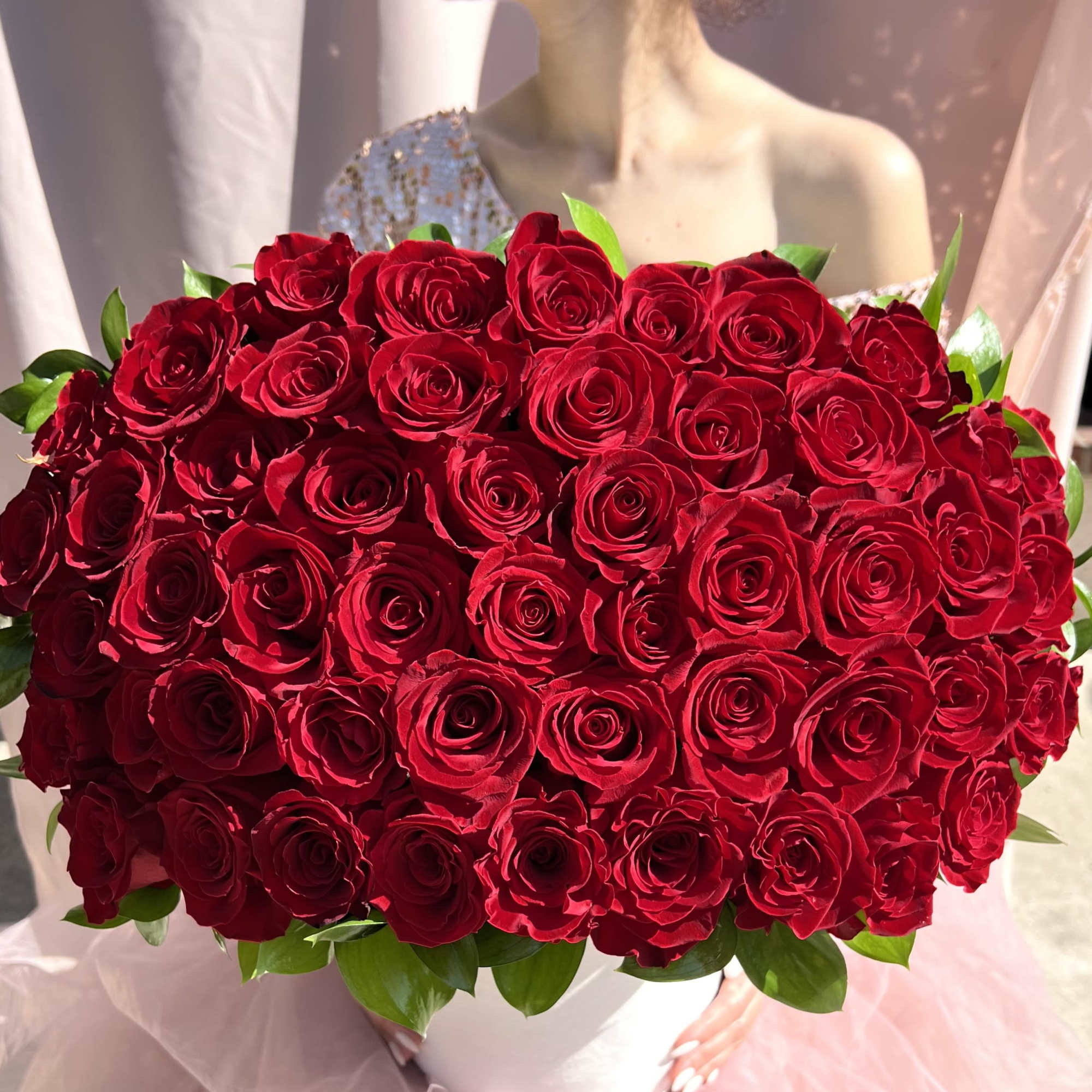 Large round arrangement of red roses in a white box held by a woman in a pink tulle skirt.