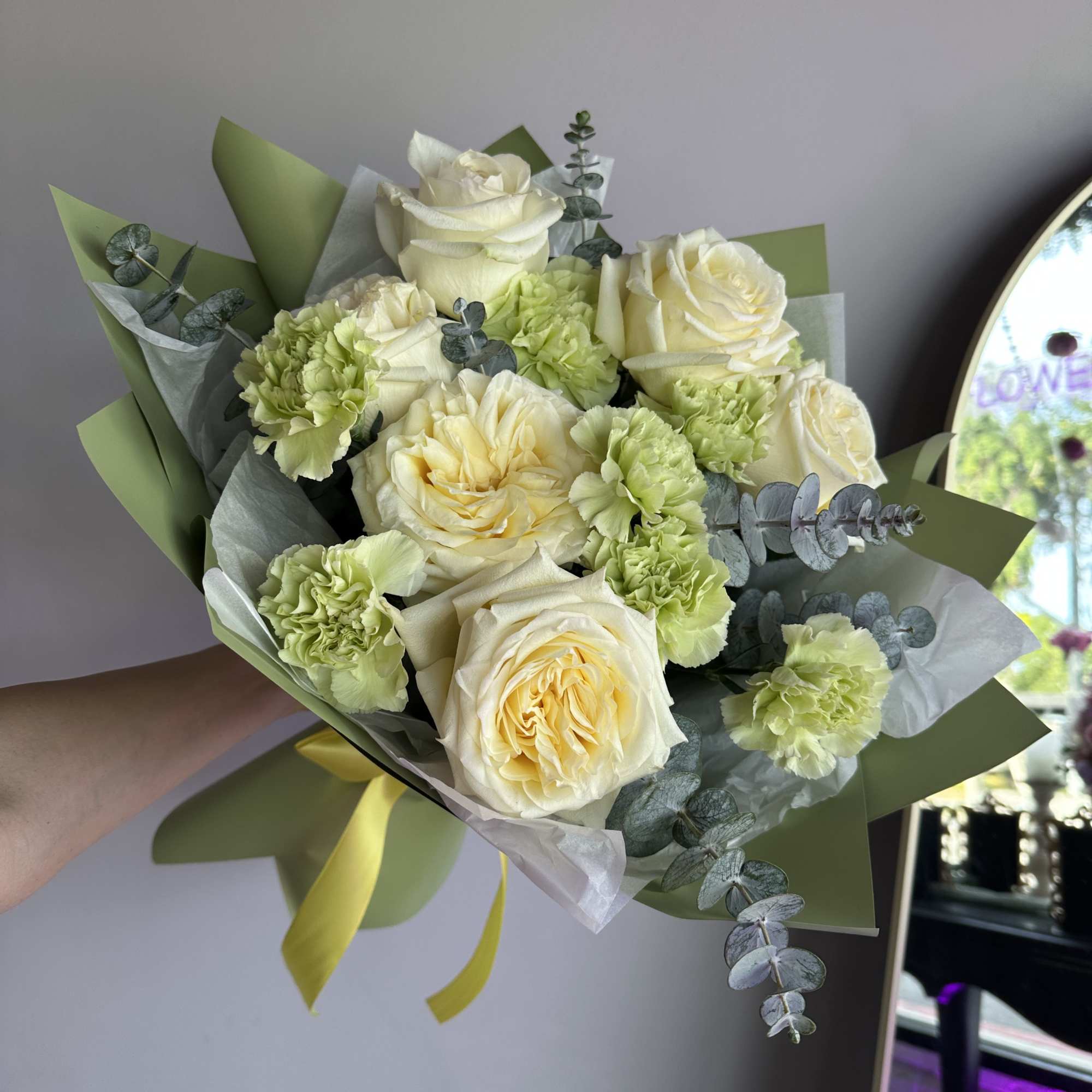 Hand-tied bouquet of pale yellow roses and light green carnations wrapped in green paper with a yellow ribbon.