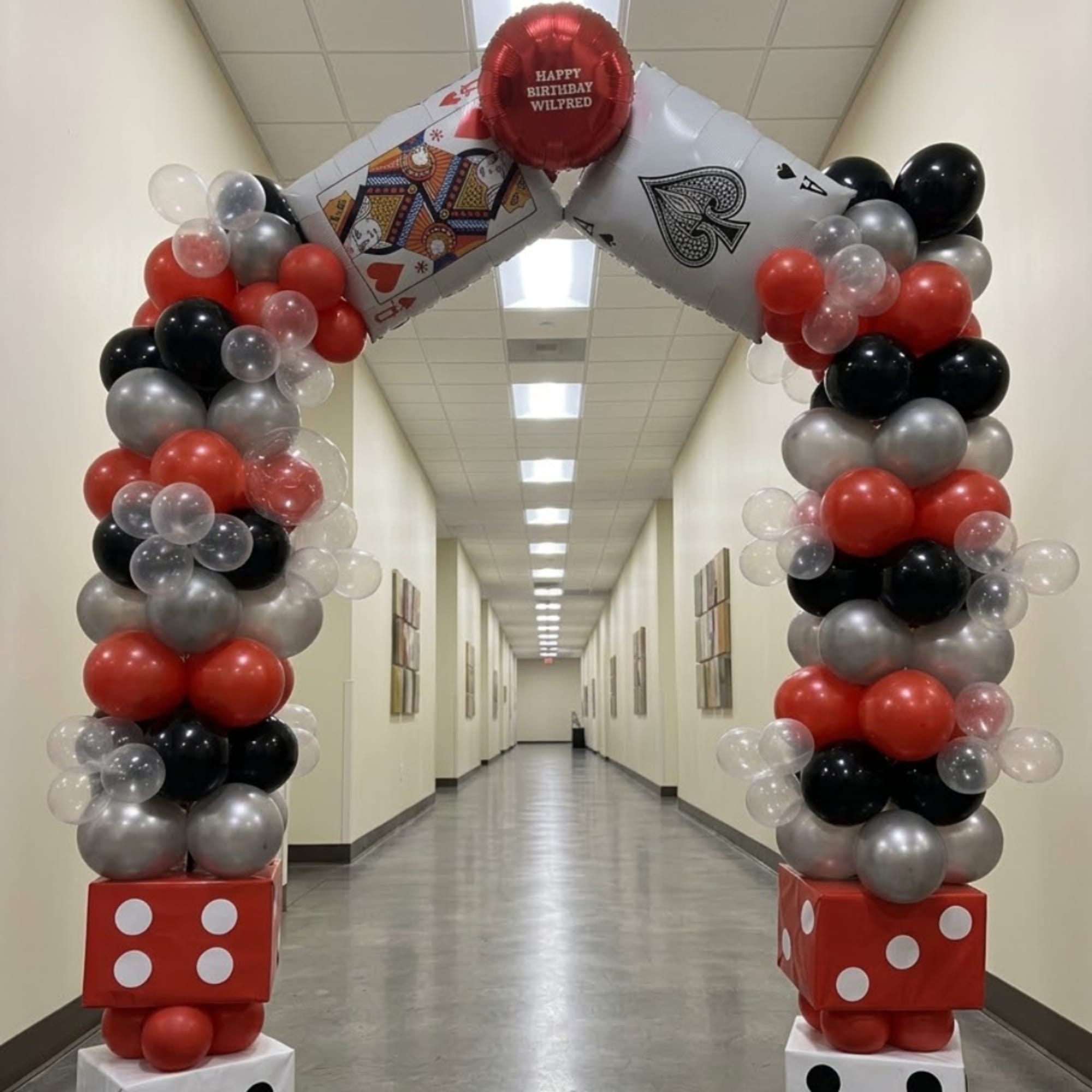 Balloon arch with playing-card decorations and a birthday balloon in a hallway