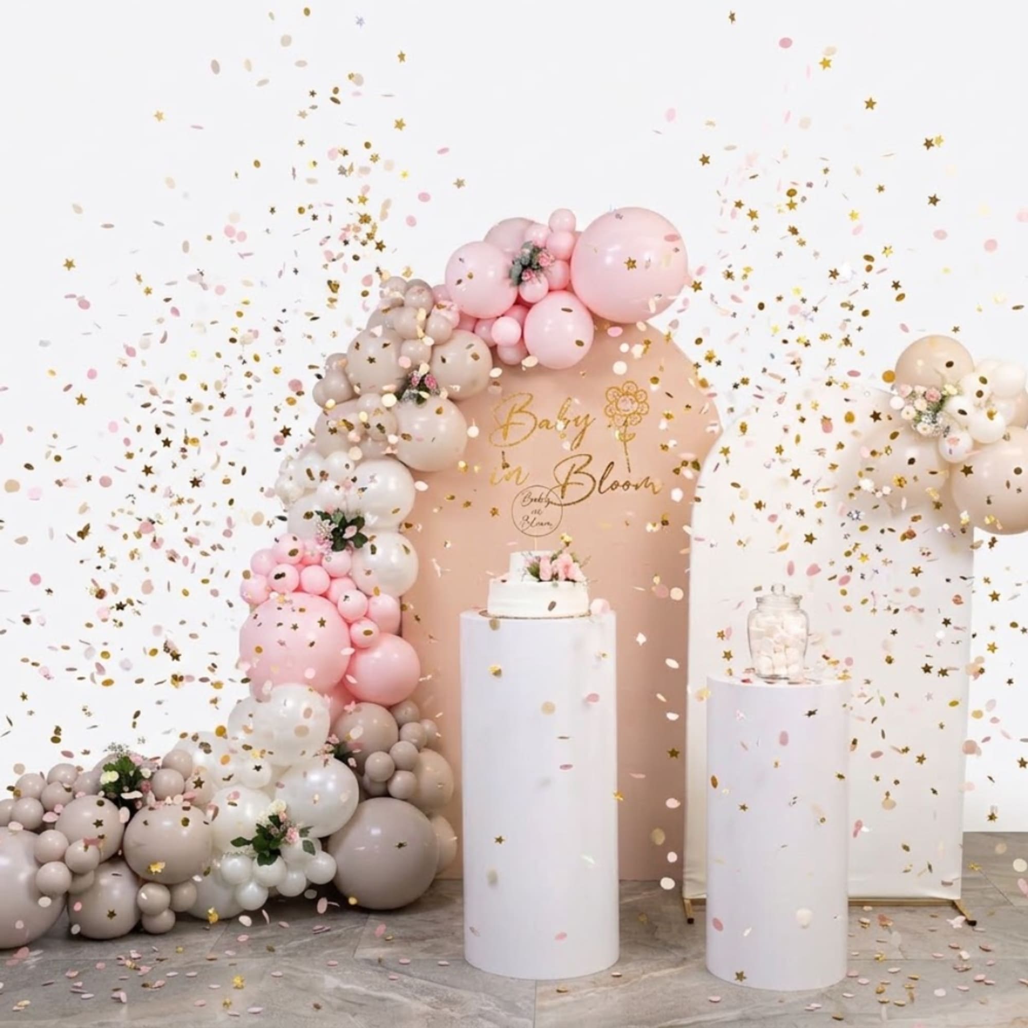 Pink and beige balloon arch with a baby shower backdrop and confetti
