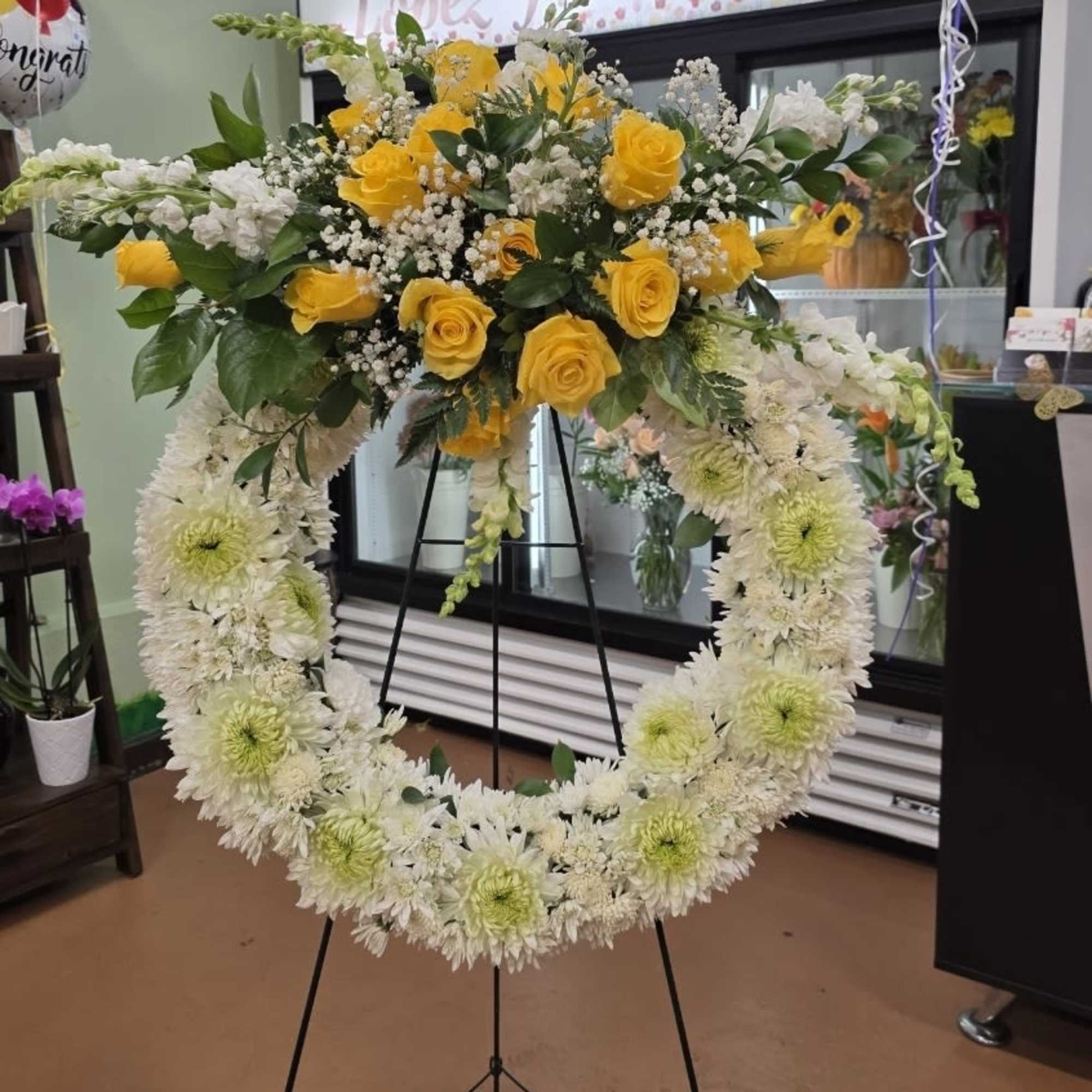 Large white chrysanthemum wreath on an easel with yellow roses and white flowers clustered at the top.