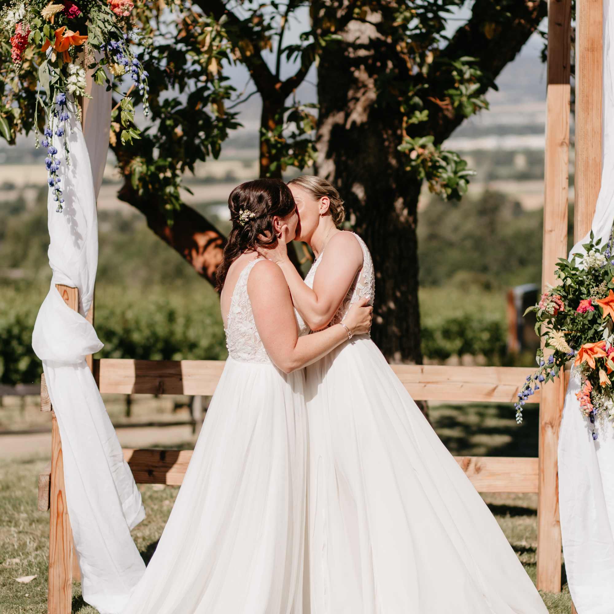 Two brides in white dresses kiss beneath a floral wedding arch.