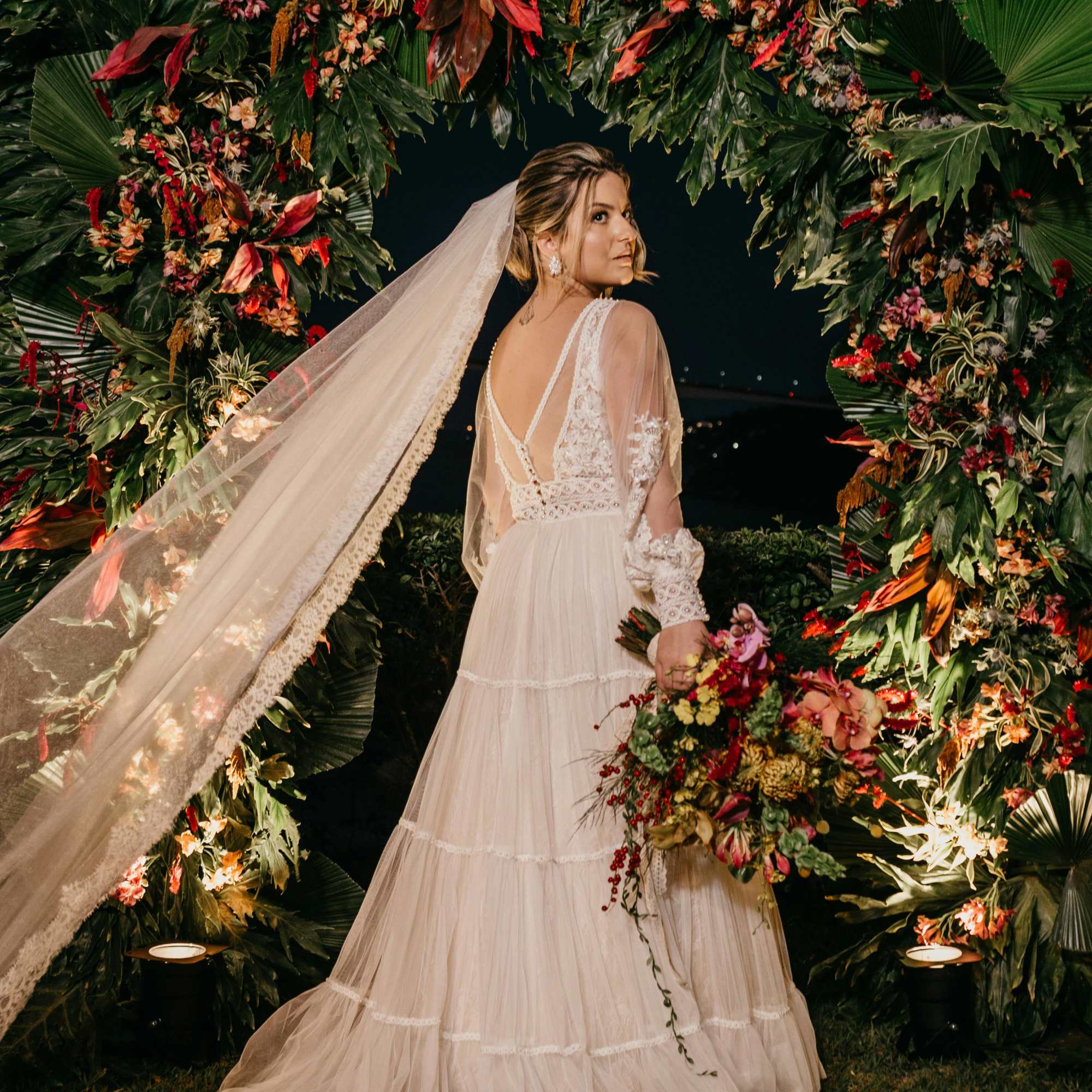 Bride in a lace gown holding a colorful bouquet under a floral arch