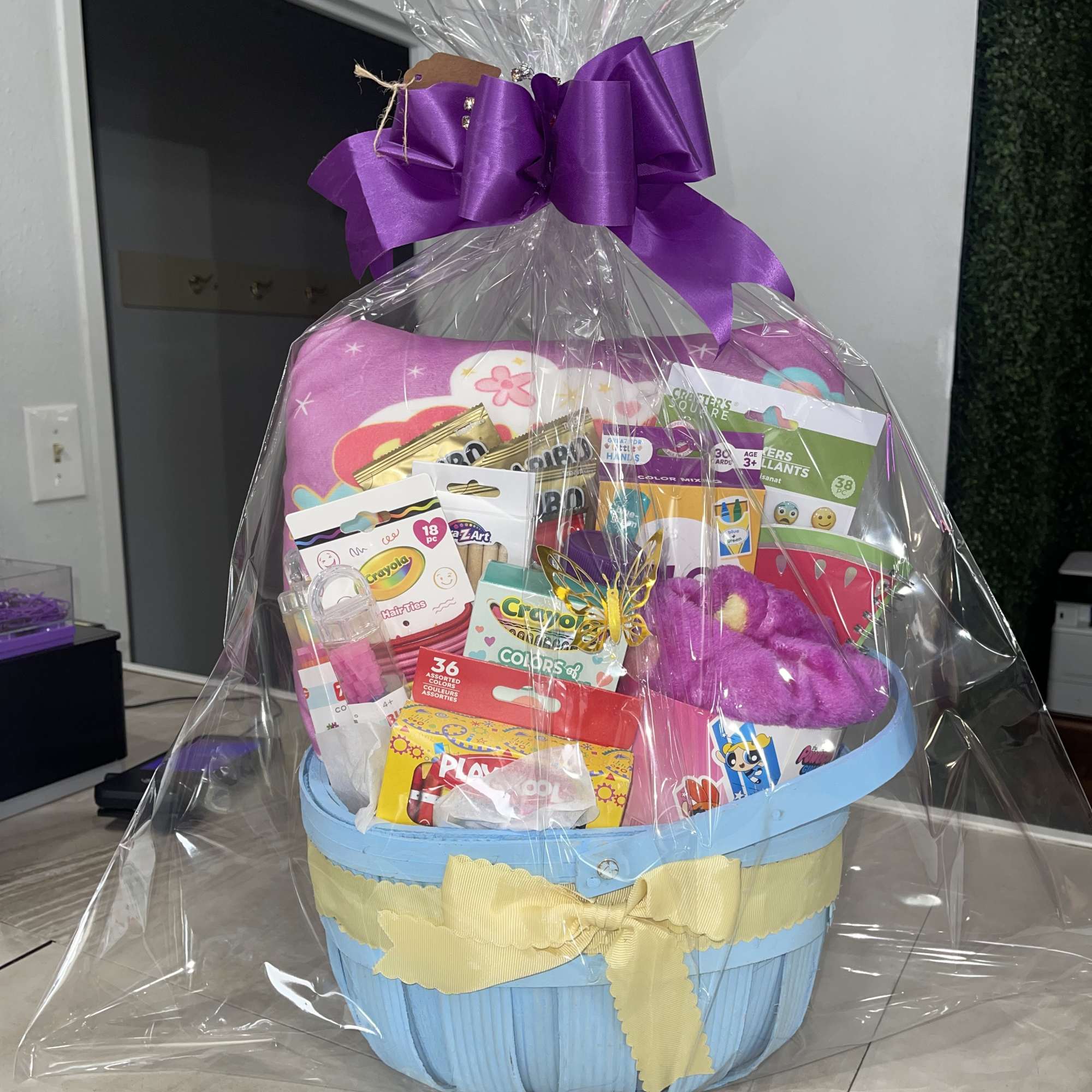 Gift basket with a purple bow and assorted craft supplies wrapped in clear cellophane