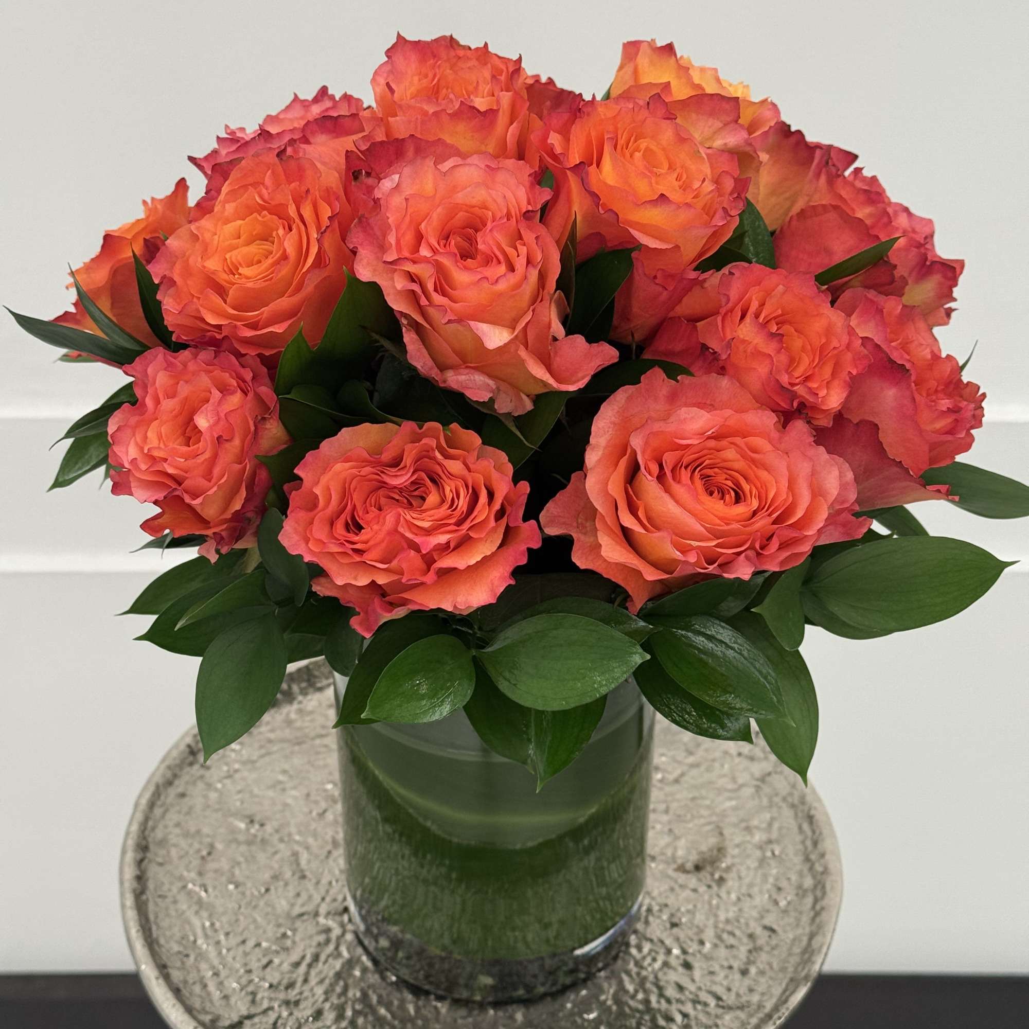 Orange roses arranged in a glass vase with dark green foliage