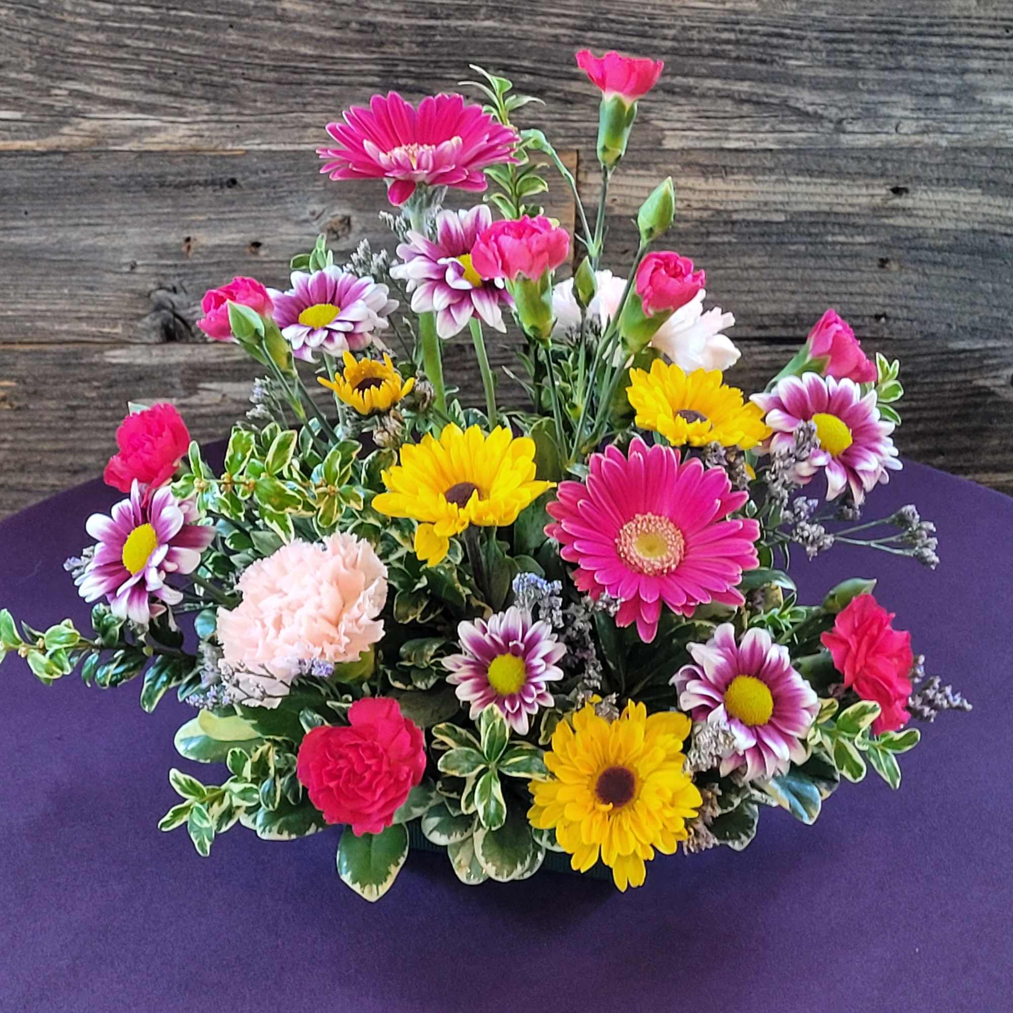 Low mixed arrangement of pink gerberas, yellow daisies, carnations and mums in a small bowl on a purple table.