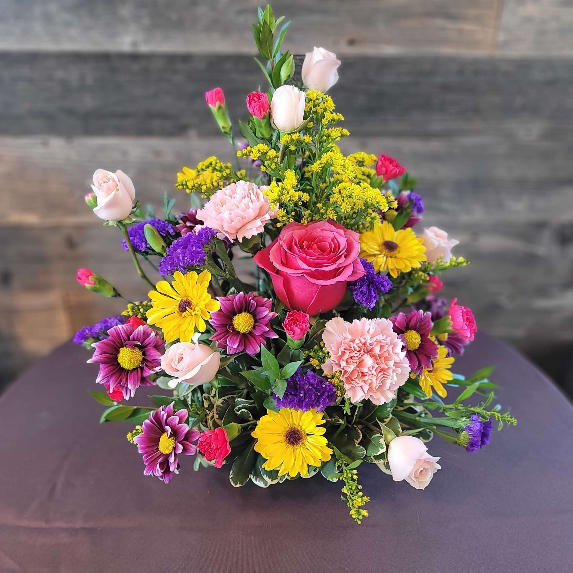 Colorful mixed arrangement with pink roses, carnations, yellow daisies and purple flowers in a low container.