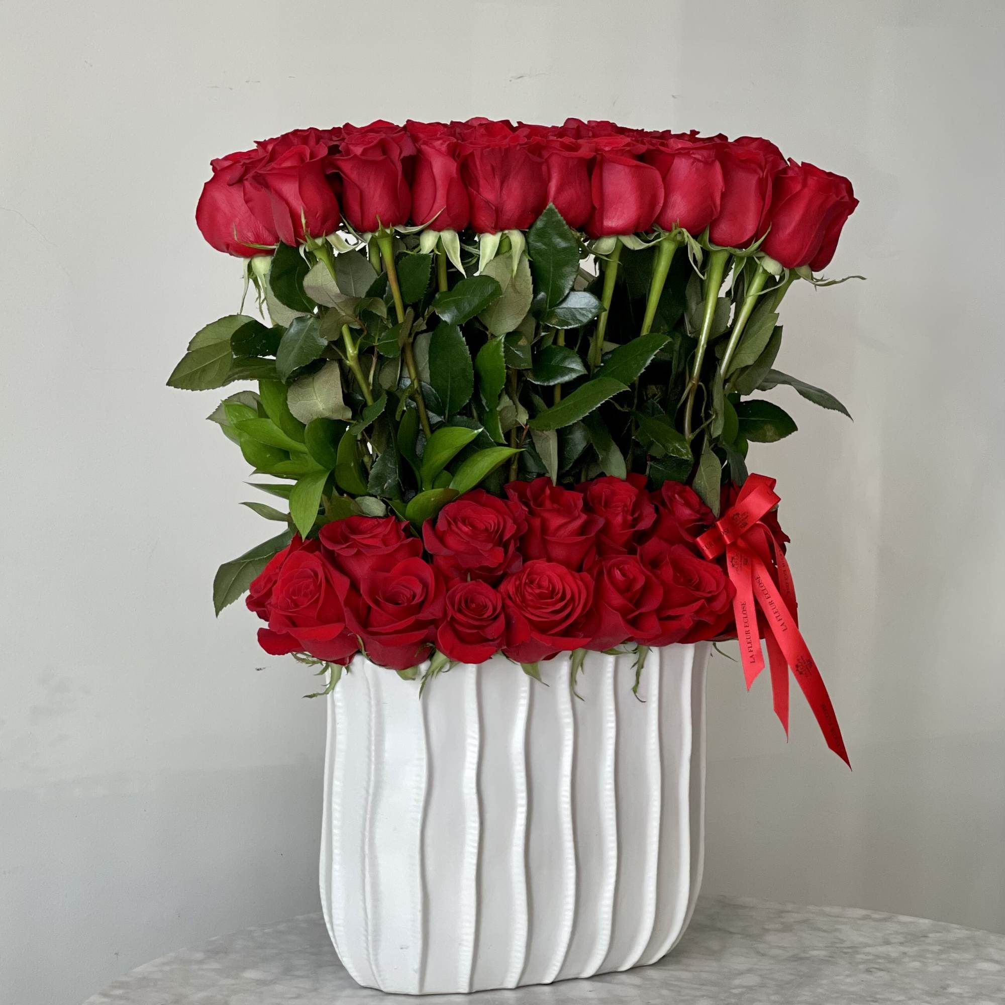Two tiers of red roses arranged in a white vase with a red ribbon