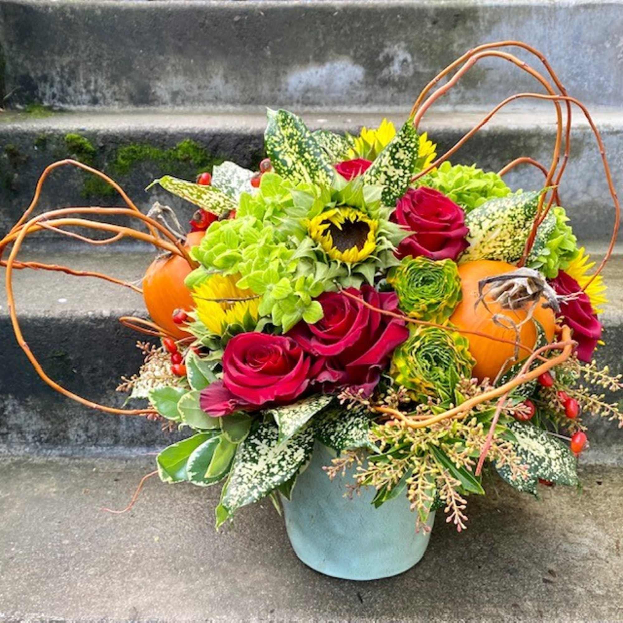 Autumn floral arrangement with roses, sunflowers, pumpkins, and curly branches in a blue container