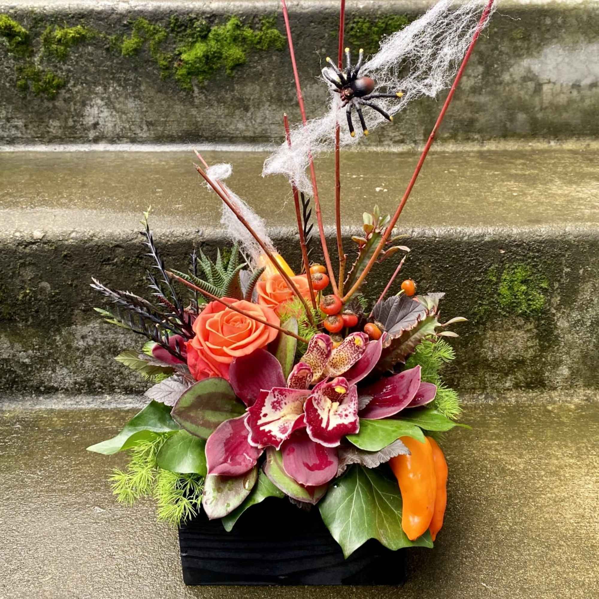 Orange roses and burgundy orchids in a black container with Halloween spider decor