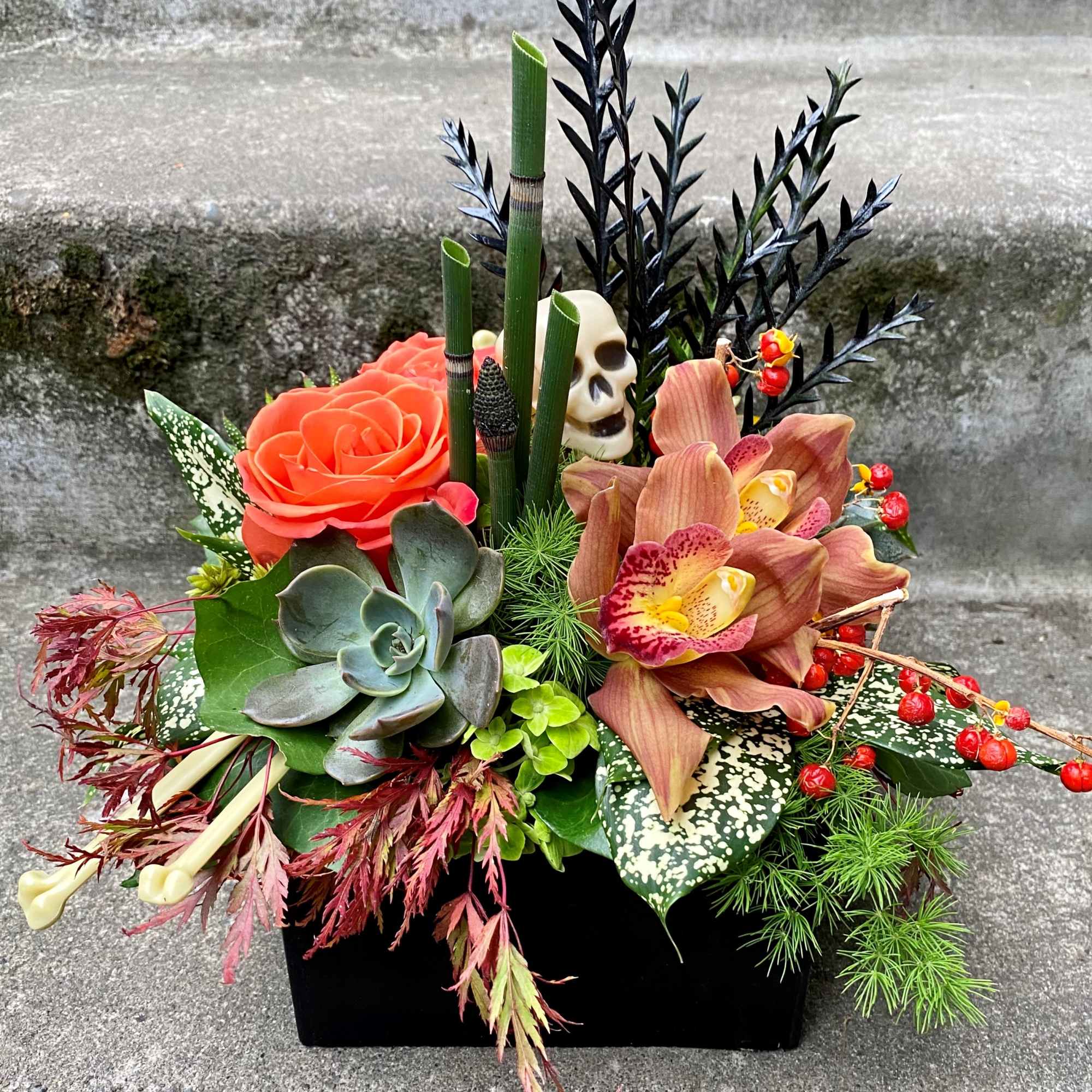 Halloween floral arrangement with orange roses, orchids, and a skull accent in a black container