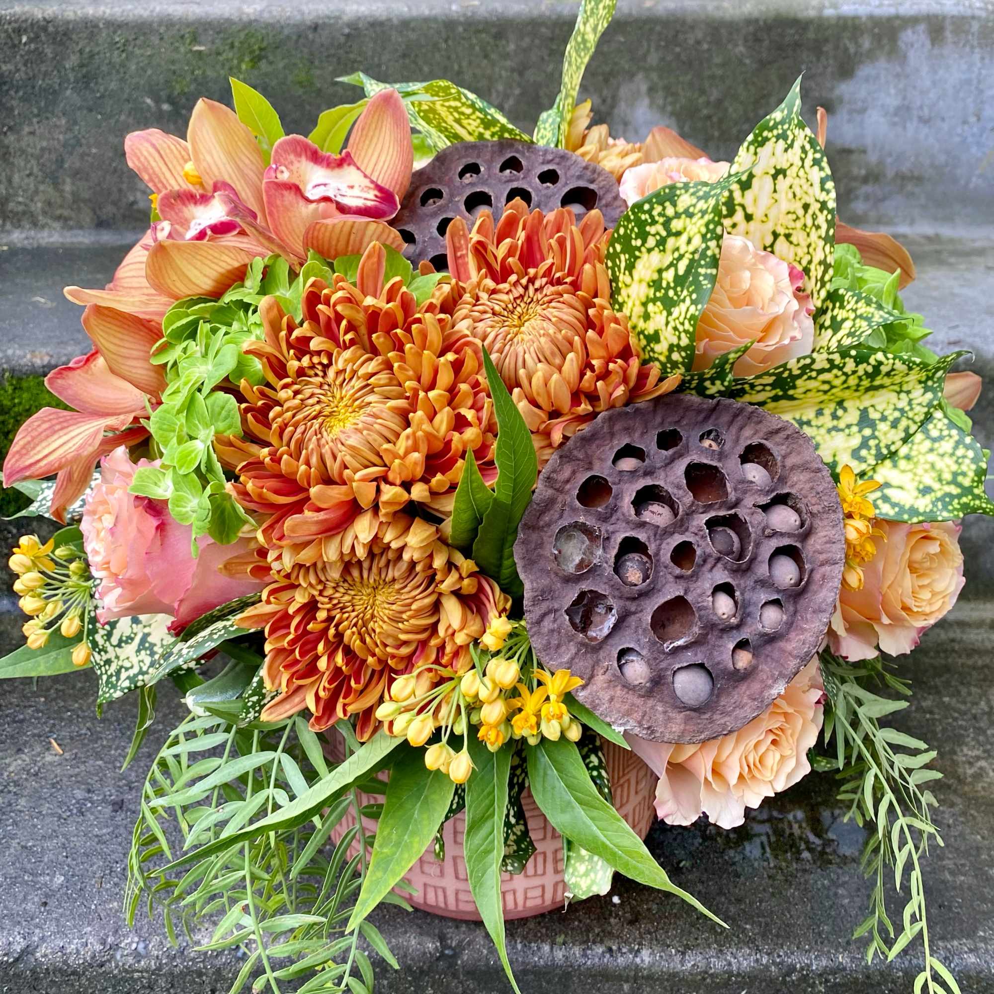 Orange and peach floral arrangement in a pink vase with lotus pods