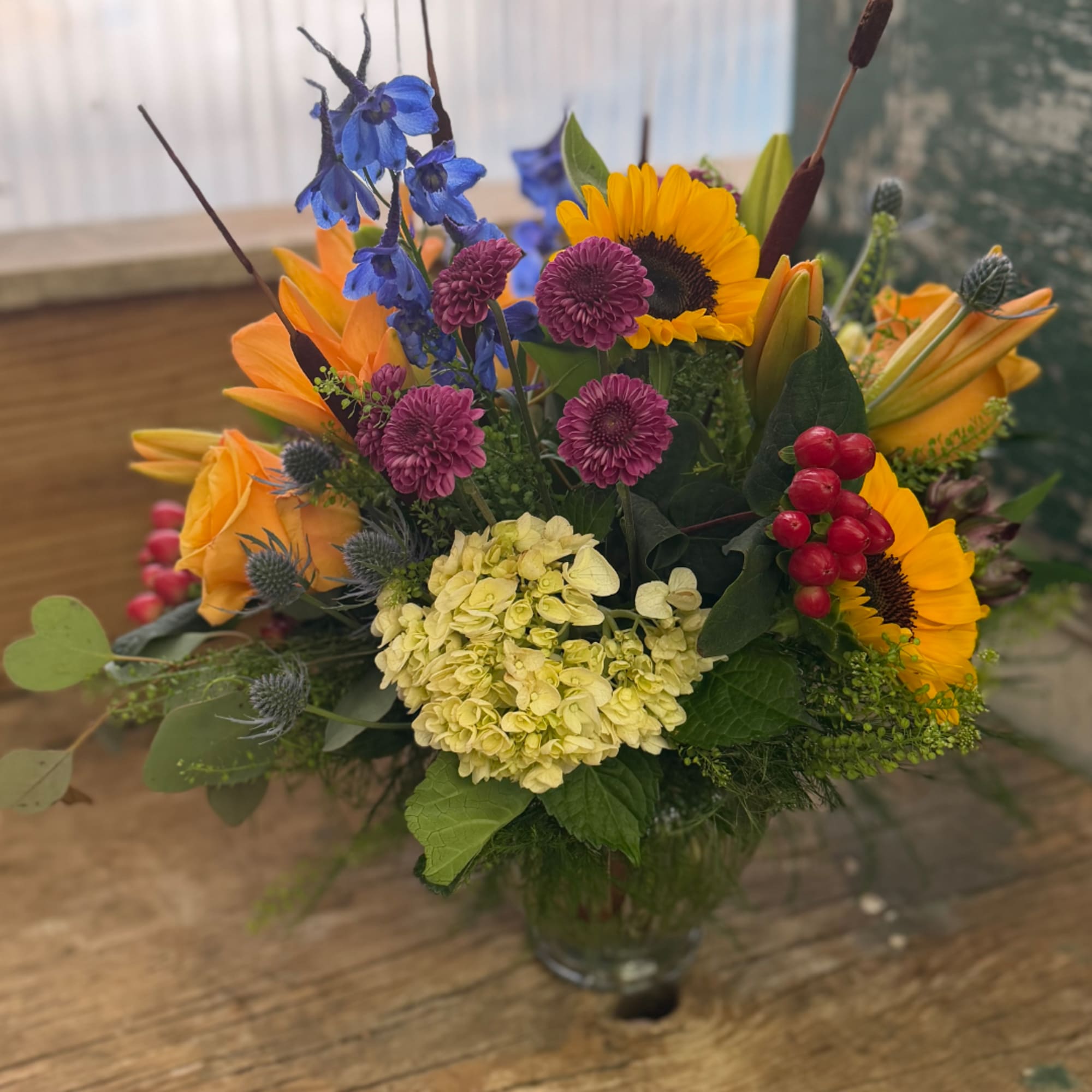 Mixed bouquet with sunflowers, orange roses, blue delphinium, and hydrangea in a glass vase