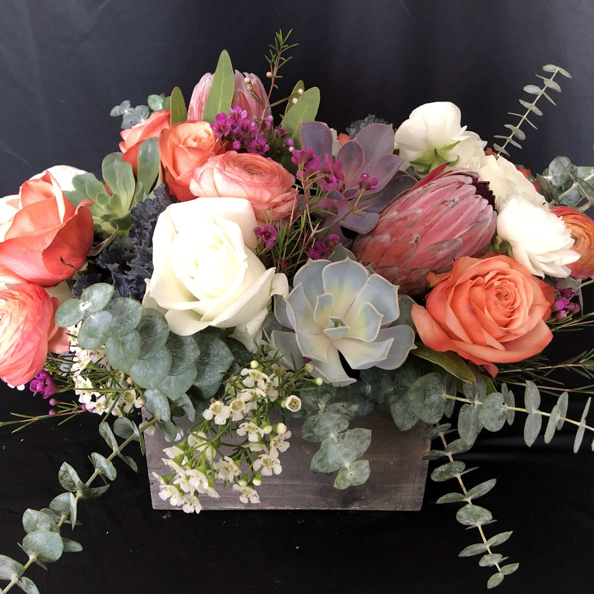 Low wooden box arrangement with orange and white roses, protea, succulents, and small accent flowers.