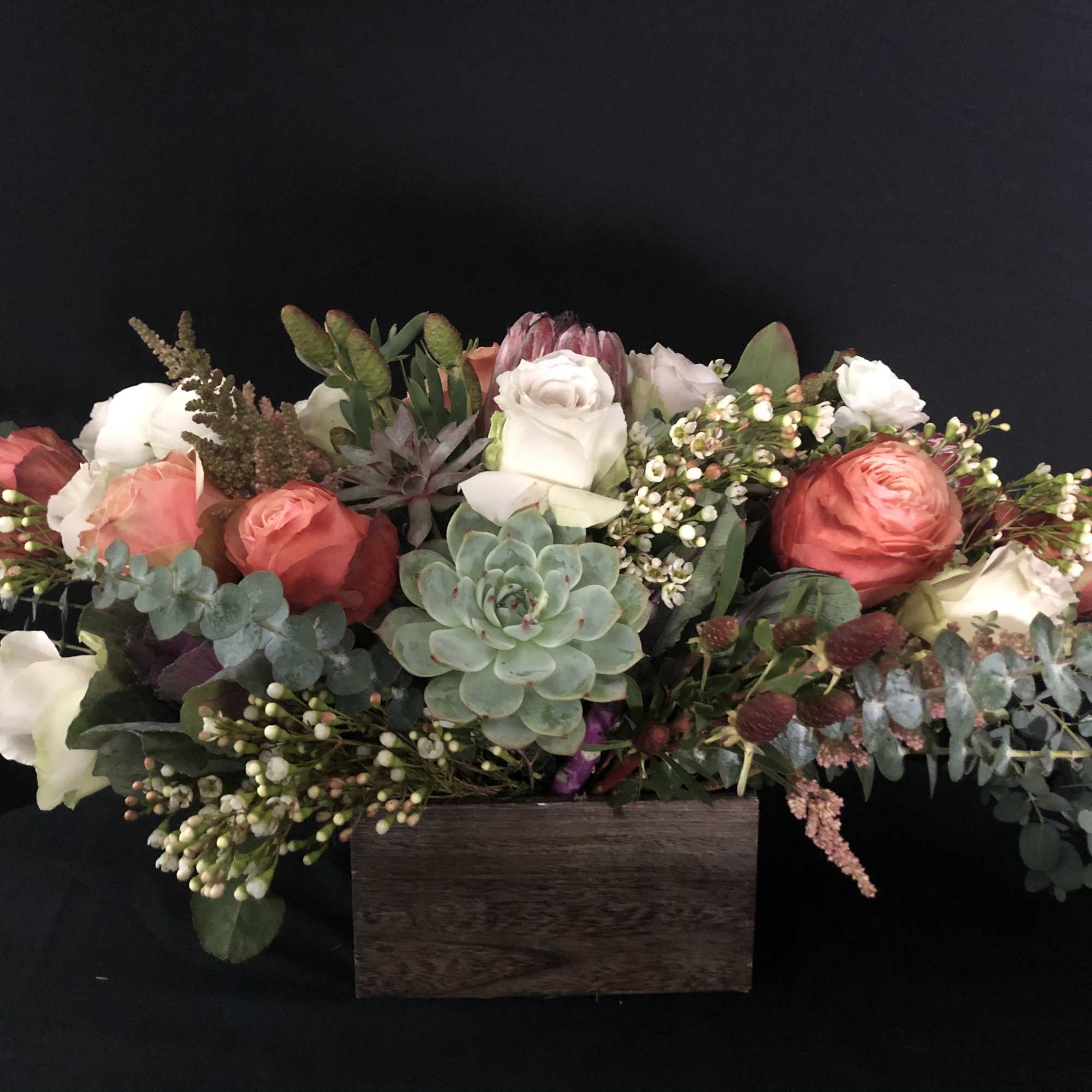 Low arrangement of coral and white roses with succulents in a rectangular wooden box.