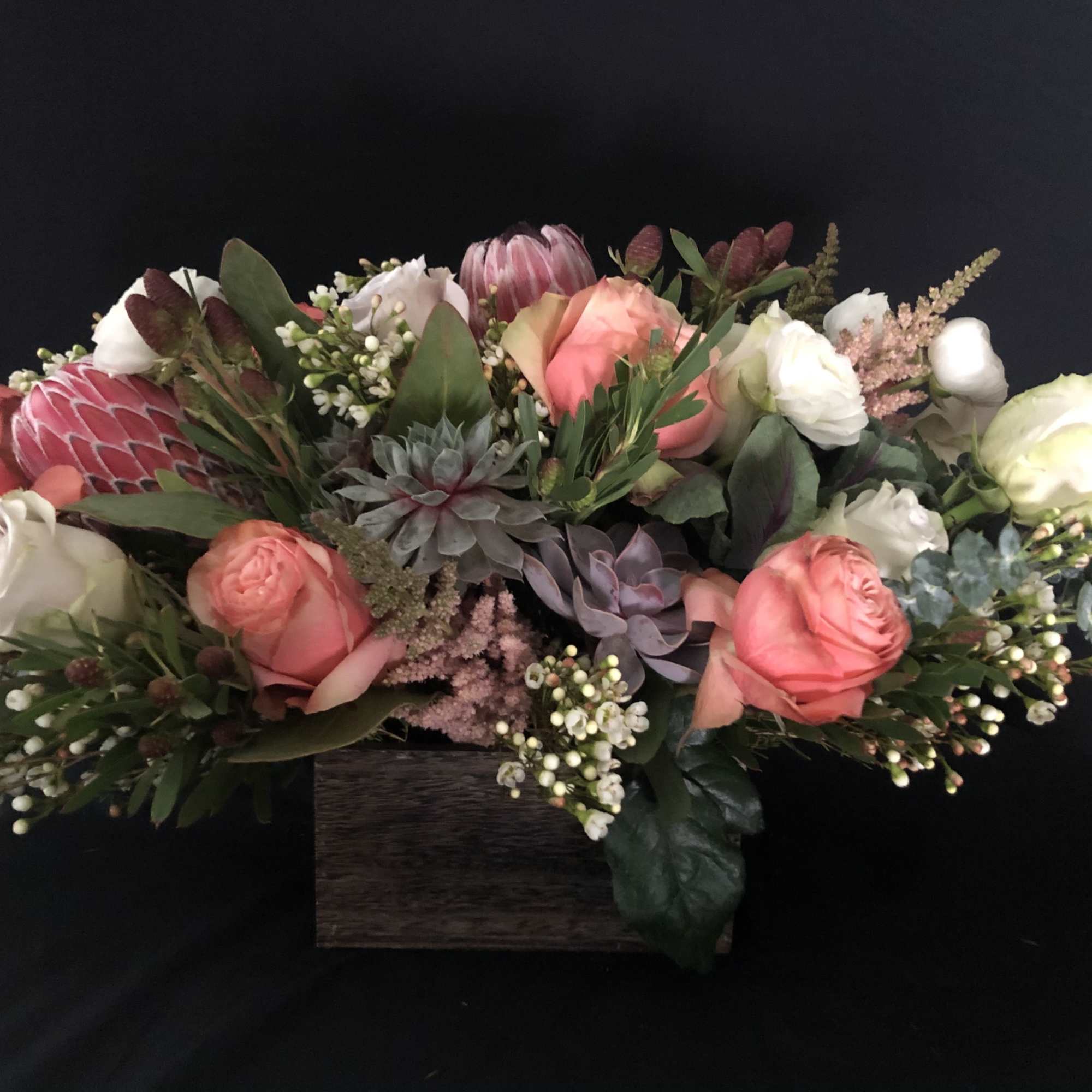 Low centerpiece of peach and white roses with protea and succulents in a dark wooden box