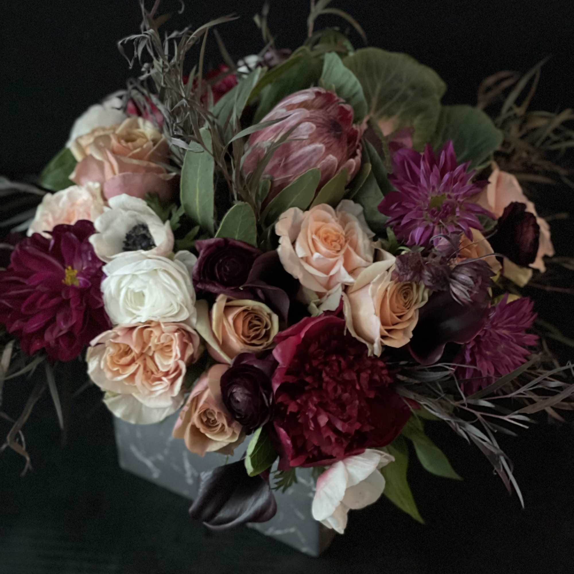 Bouquet of roses, protea, and dark blooms in a gray box
