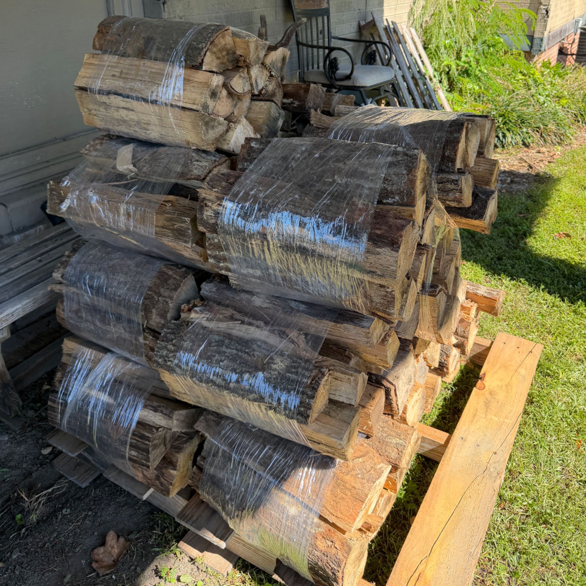 Stacked firewood wrapped in plastic beside a porch and lawn