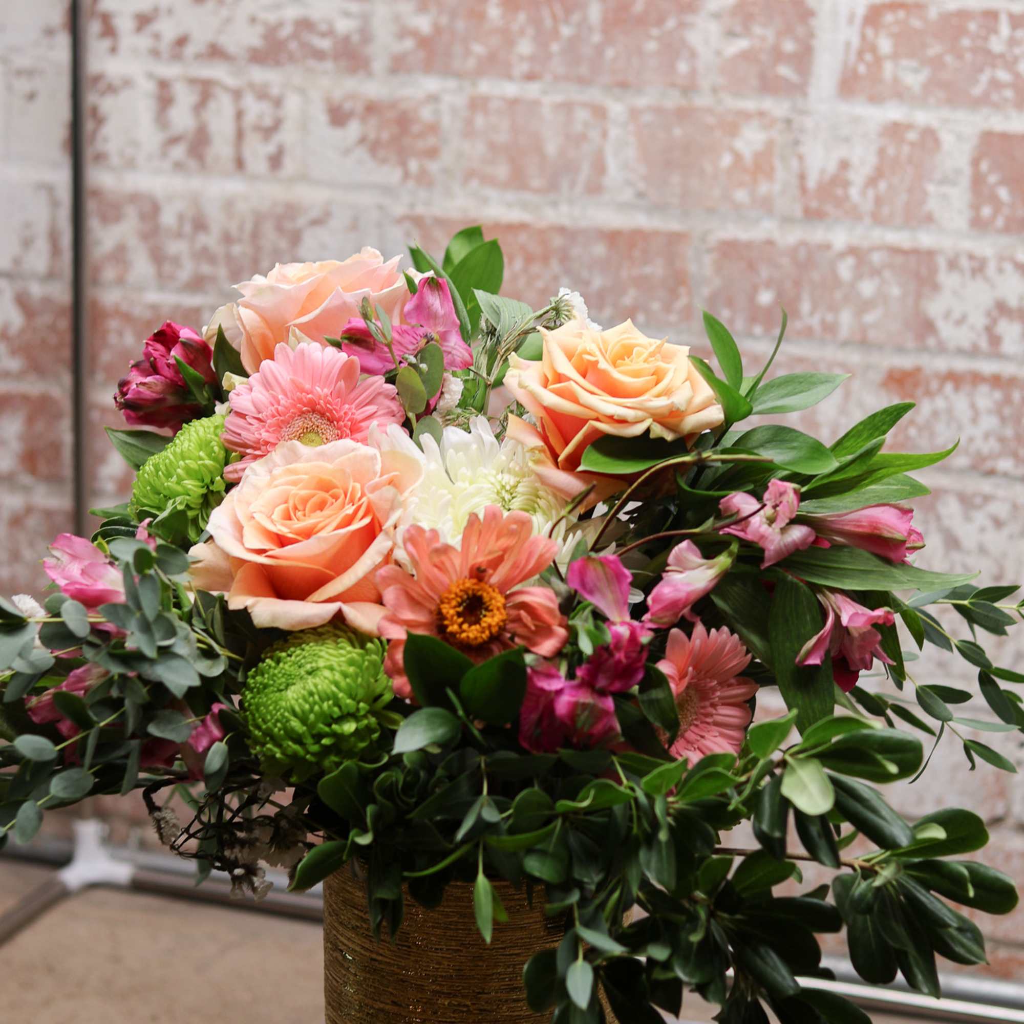 Mixed bouquet of peach roses, pink gerberas, and green mums in a gold vase