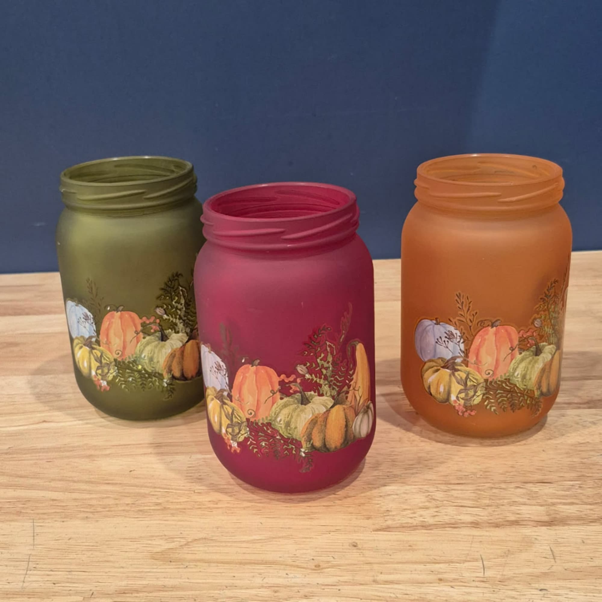 Three painted mason jars with pumpkin designs on a wooden table