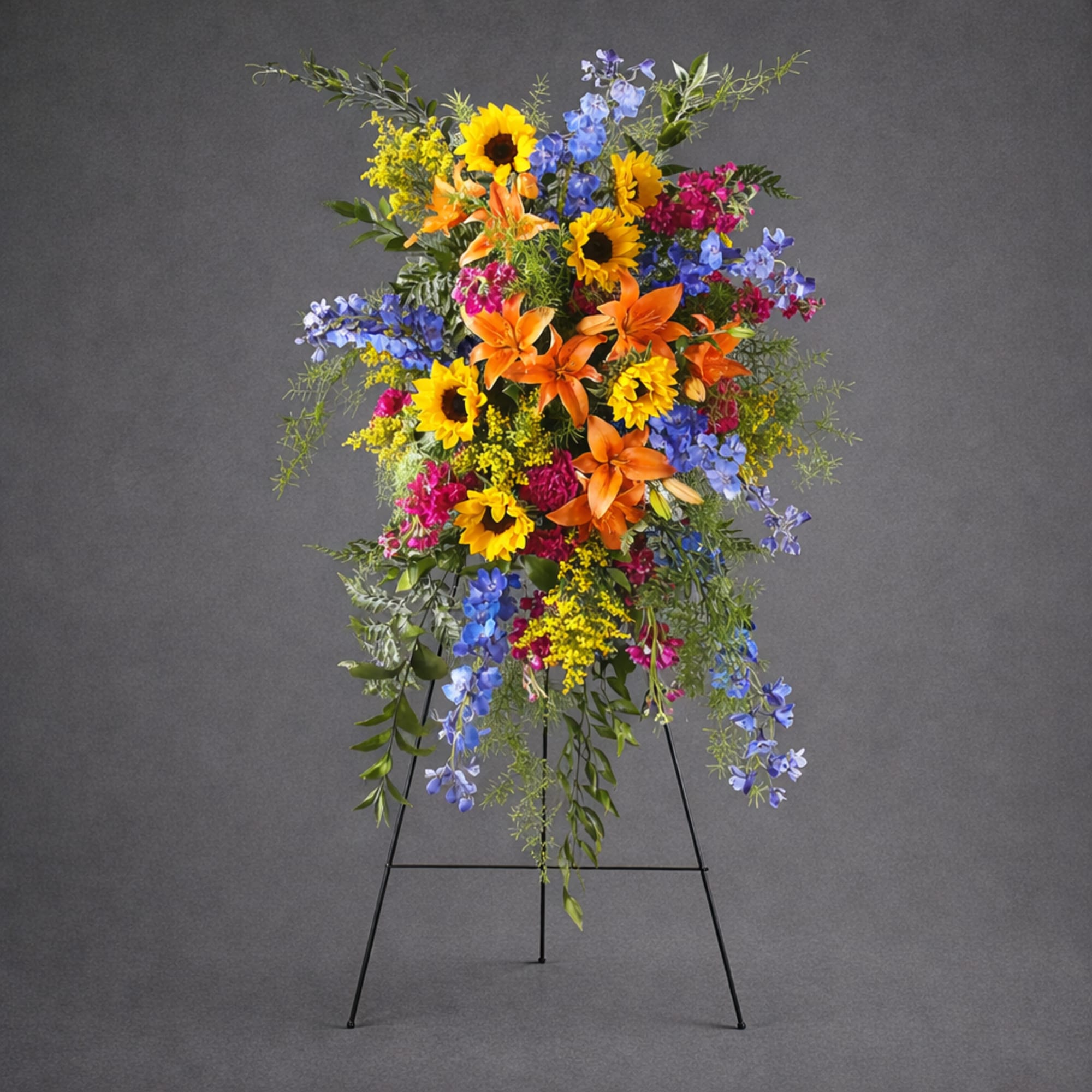 Tall standing floral spray with sunflowers, orange lilies, and blue flowers on a black easel.