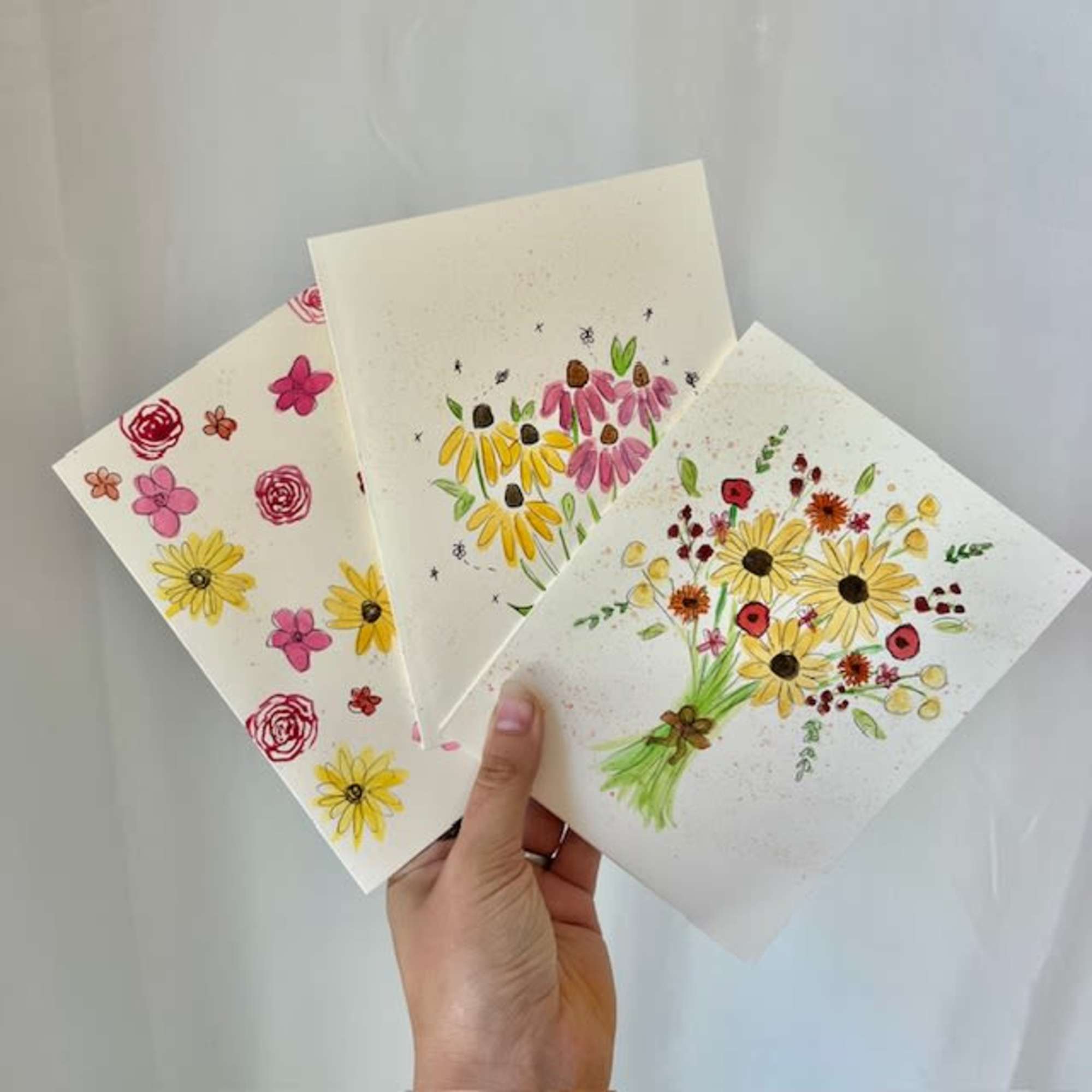 Hand holding three floral greeting cards with painted daisies and roses