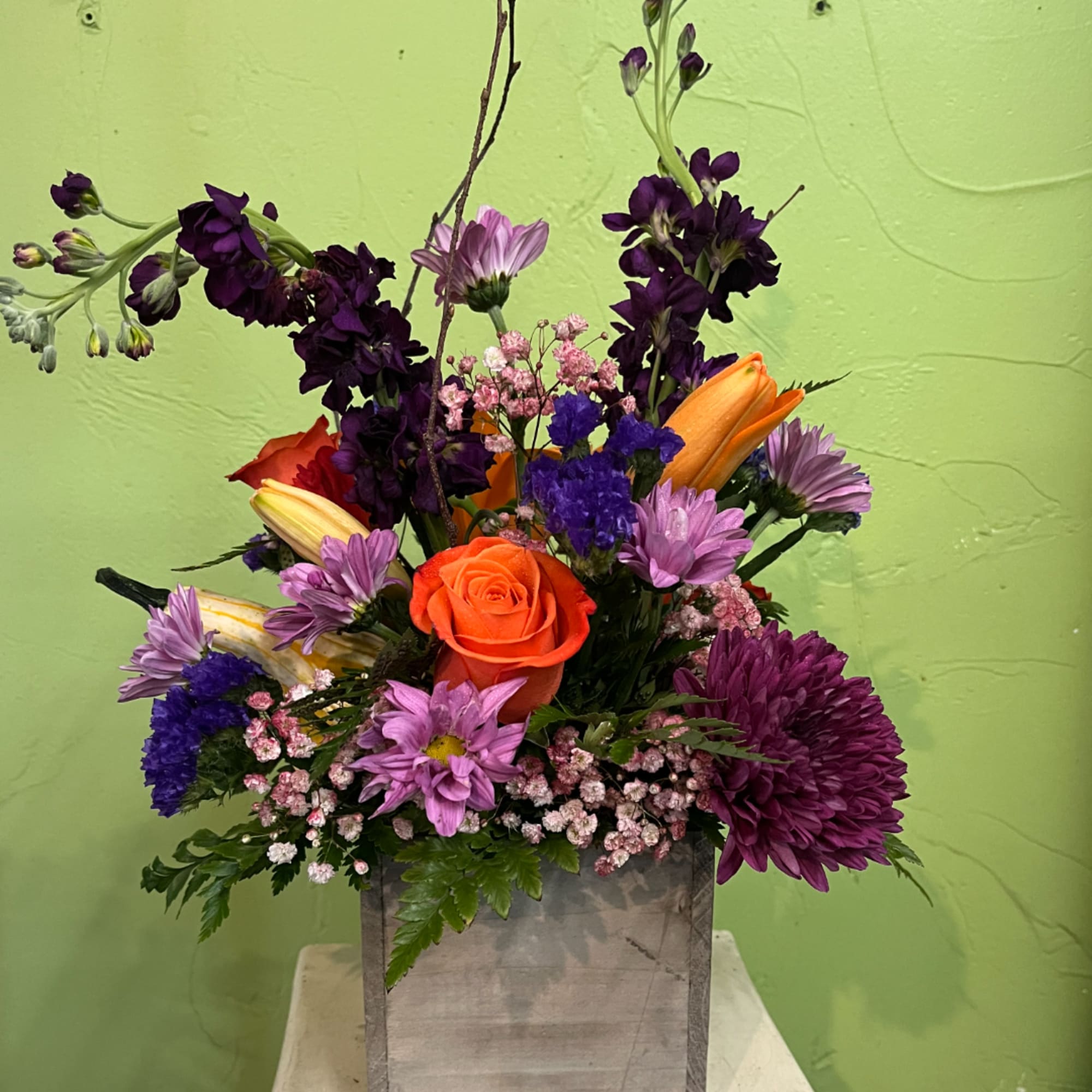 Mixed bouquet with an orange rose, lilies, and purple blooms in a gray container
