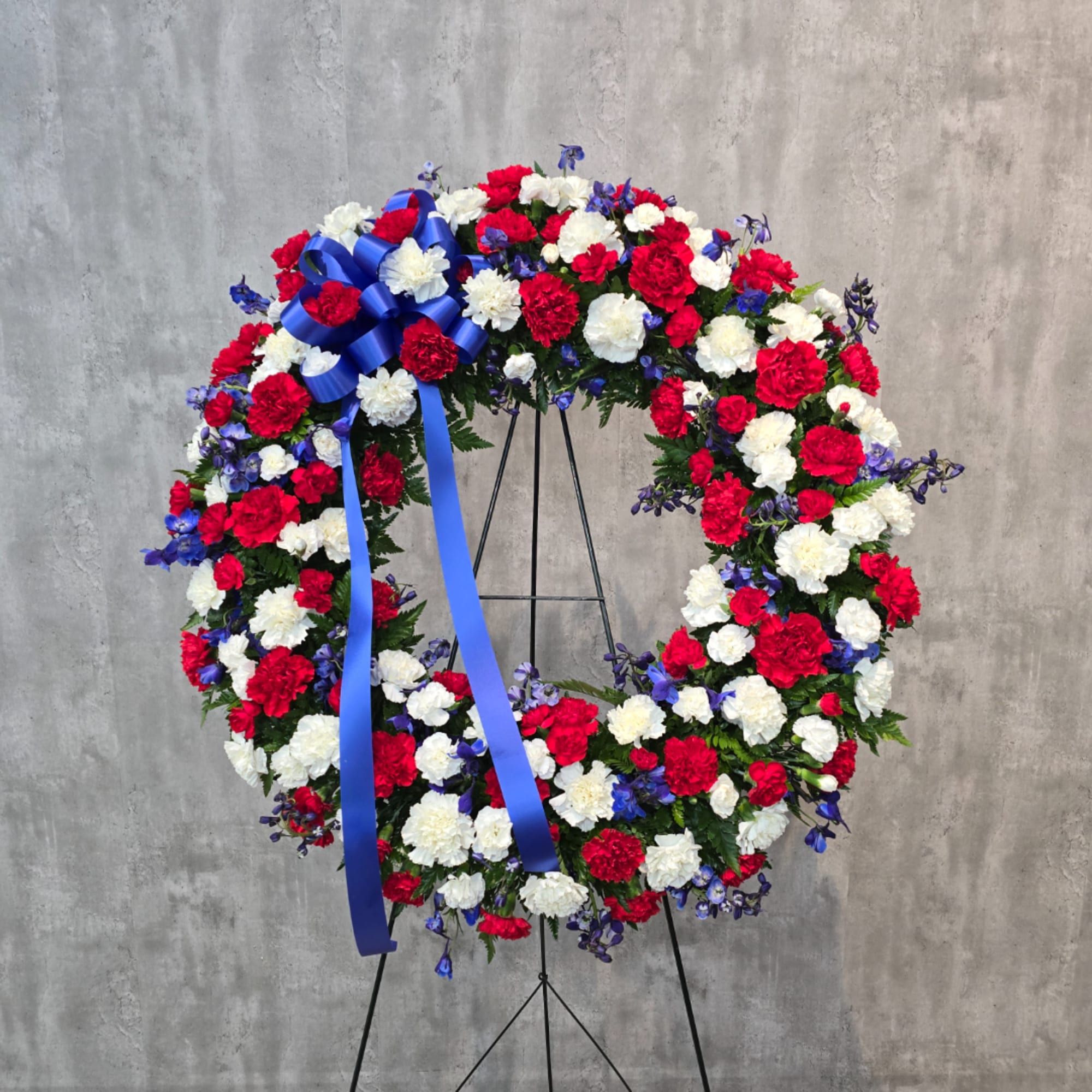 Circular wreath of red, white, and blue flowers on a stand with a blue ribbon