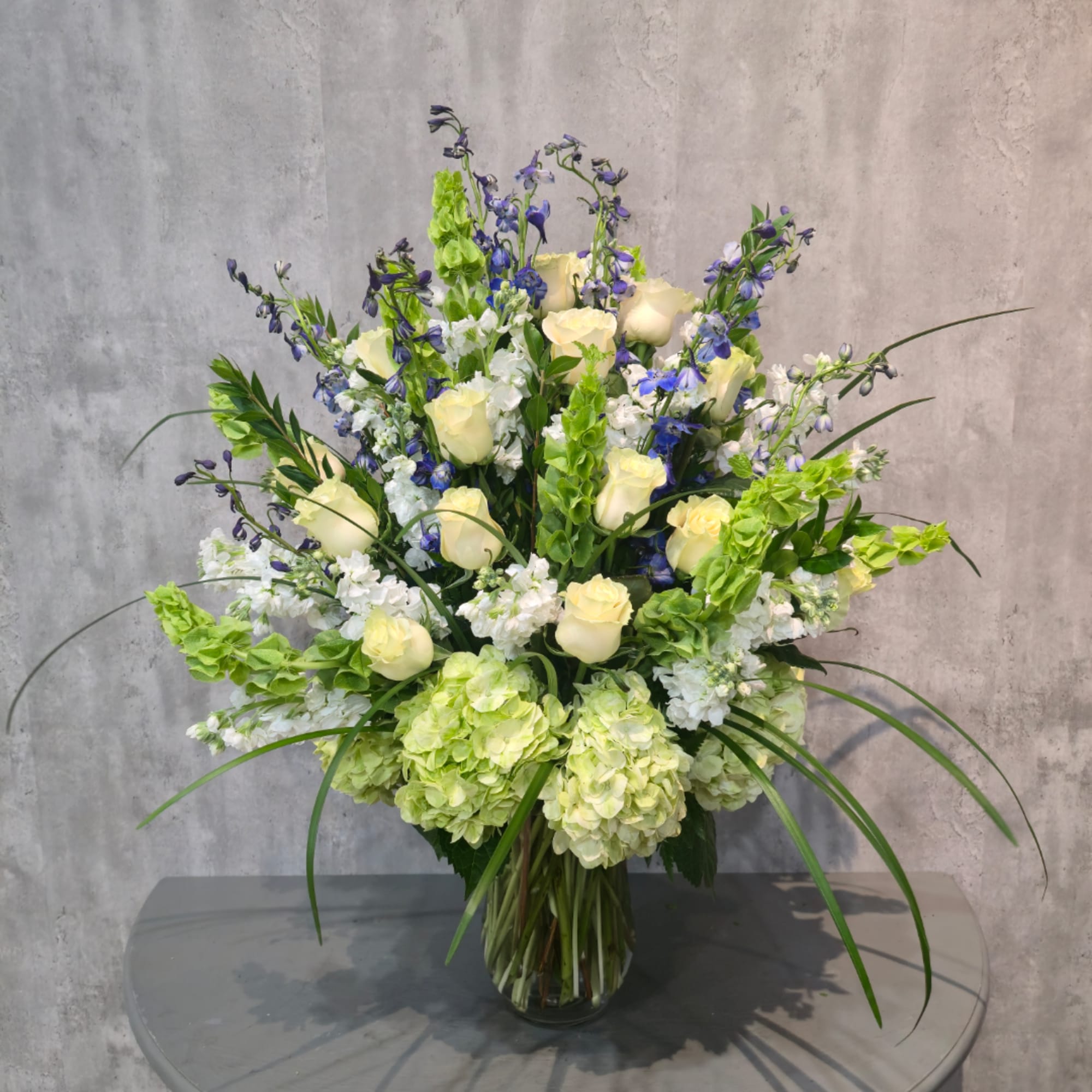 Tall bouquet of cream roses, blue flowers, and green hydrangeas in a glass vase
